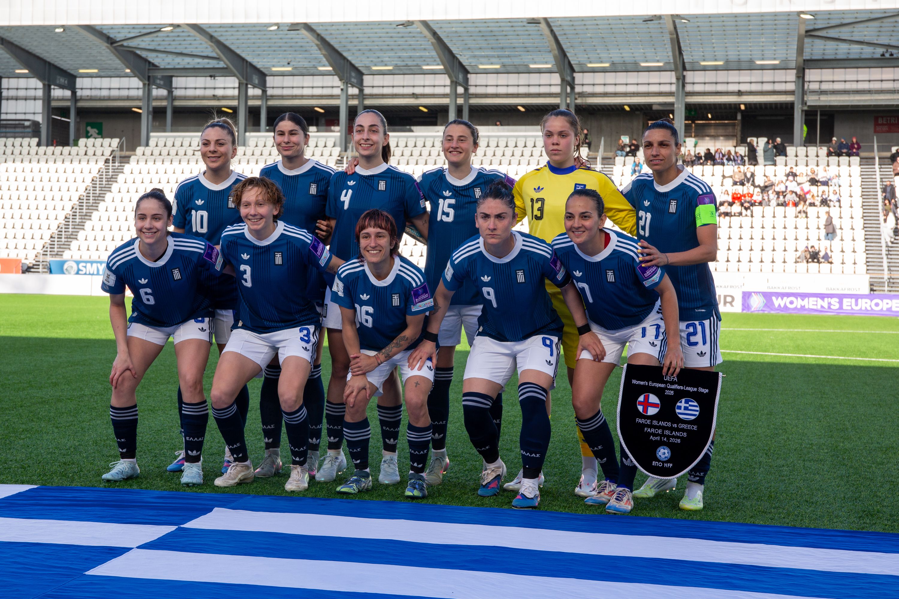 Eine Frauenfußballmannschaft posiert in blauen Trikots auf einem Spielfeld, mit Trainerin und Flagge im Vordergrund.