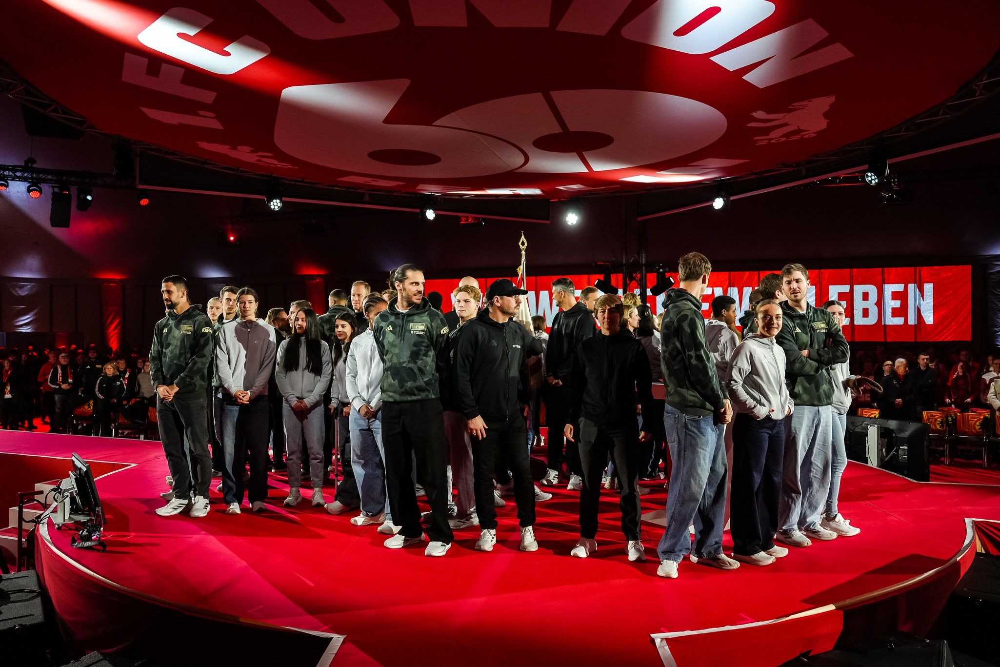 Eine Gruppe von Menschen steht auf einer Bühne, umgeben von Publikum und dekorativen Bannern im Zusammenhang mit einem Event.