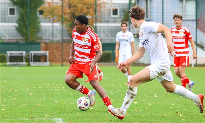 Spieler in roten und weißen Trikots stehen auf einem Fußballfeld. Ein Spieler dribbelt den Ball, während ein anderer ihn verfolgt.