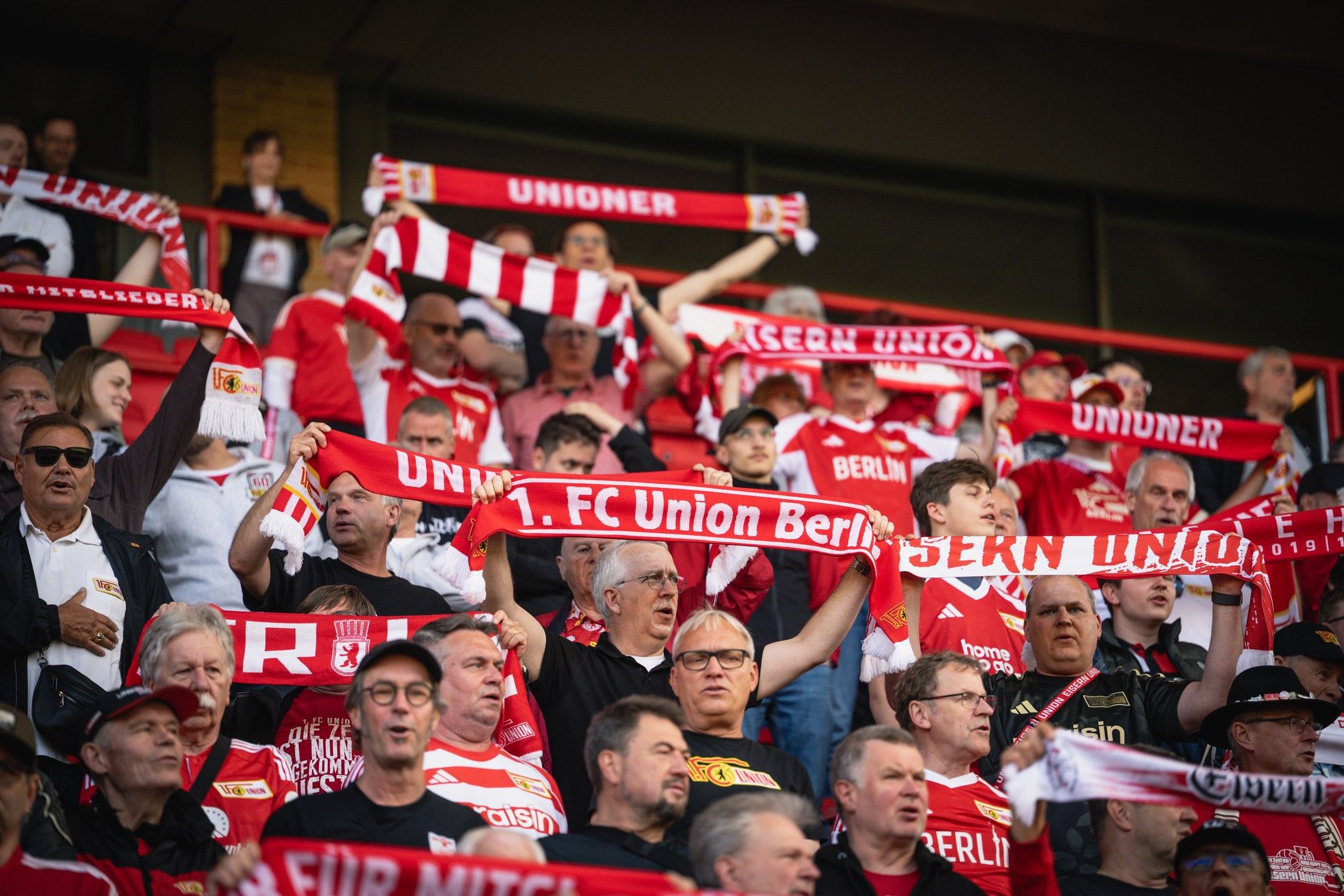 Eine Gruppe von Fans des 1. FC Union Berlin trägt rote und weiße Schals, jubelt und feuert ihr Team an.