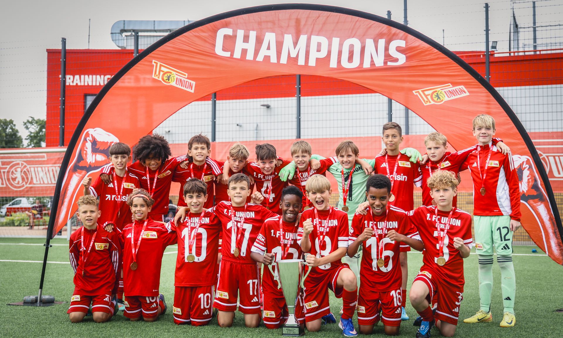 Eine Gruppe von Kindern in roten Fußballtrikots posiert stolz mit einem Pokal unter einem Champions-Banner.