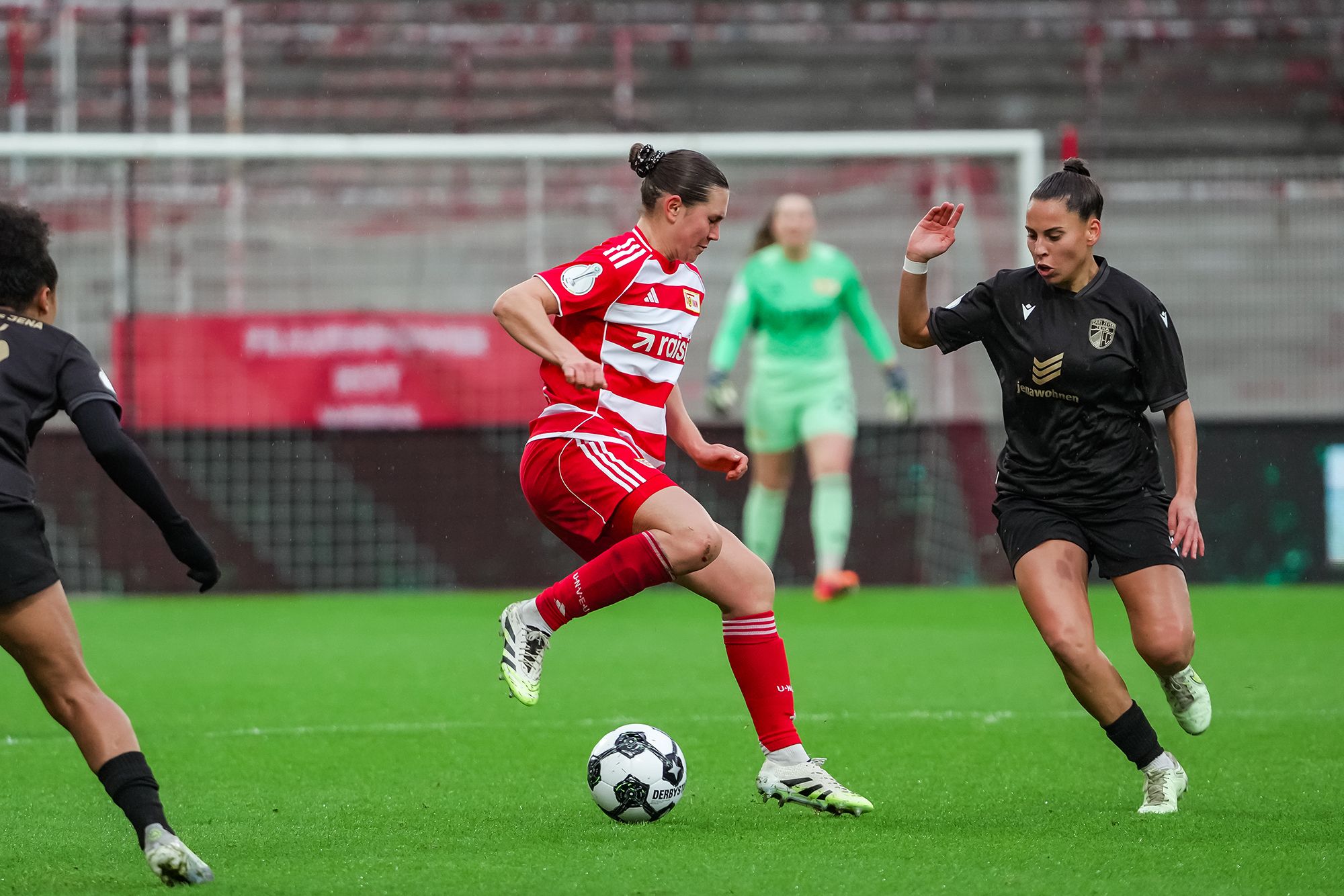Eine Fußballspielerin in rot-weißer Kleidung dribbelt den Ball, während eine Gegnerin in schwarzer Kleidung versucht, ihn zu erreichen.
