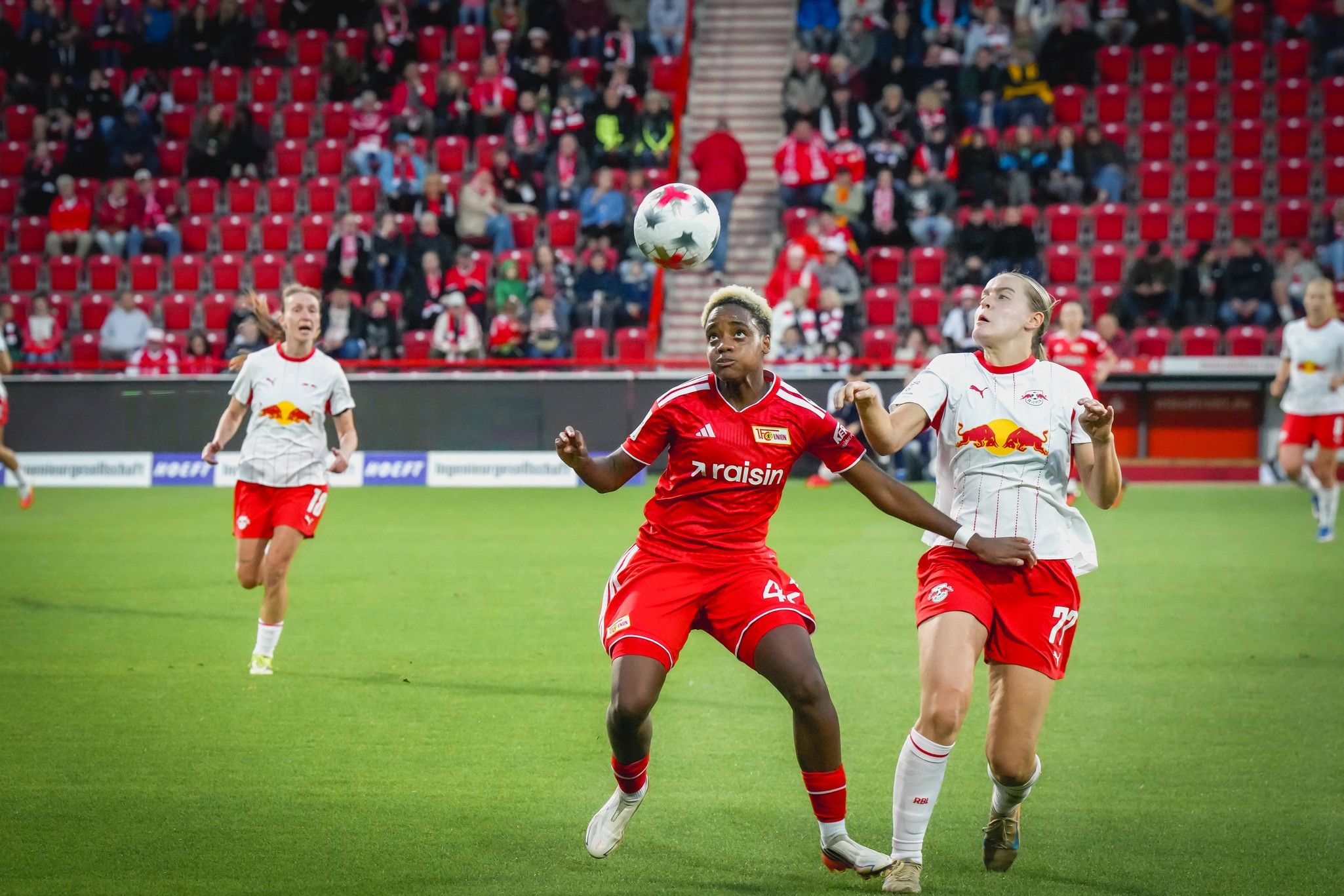 Zwei Fußballspielerinnen im Wettkampf um den Ball auf einem Fußballfeld, im Hintergrund stehen Zuschauer auf der Tribüne.