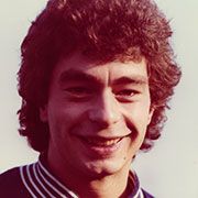 Ein lächelnder Mann mit lockigem, braunem Haar, der in einem Polohemd mit Kragen abgebildet ist.