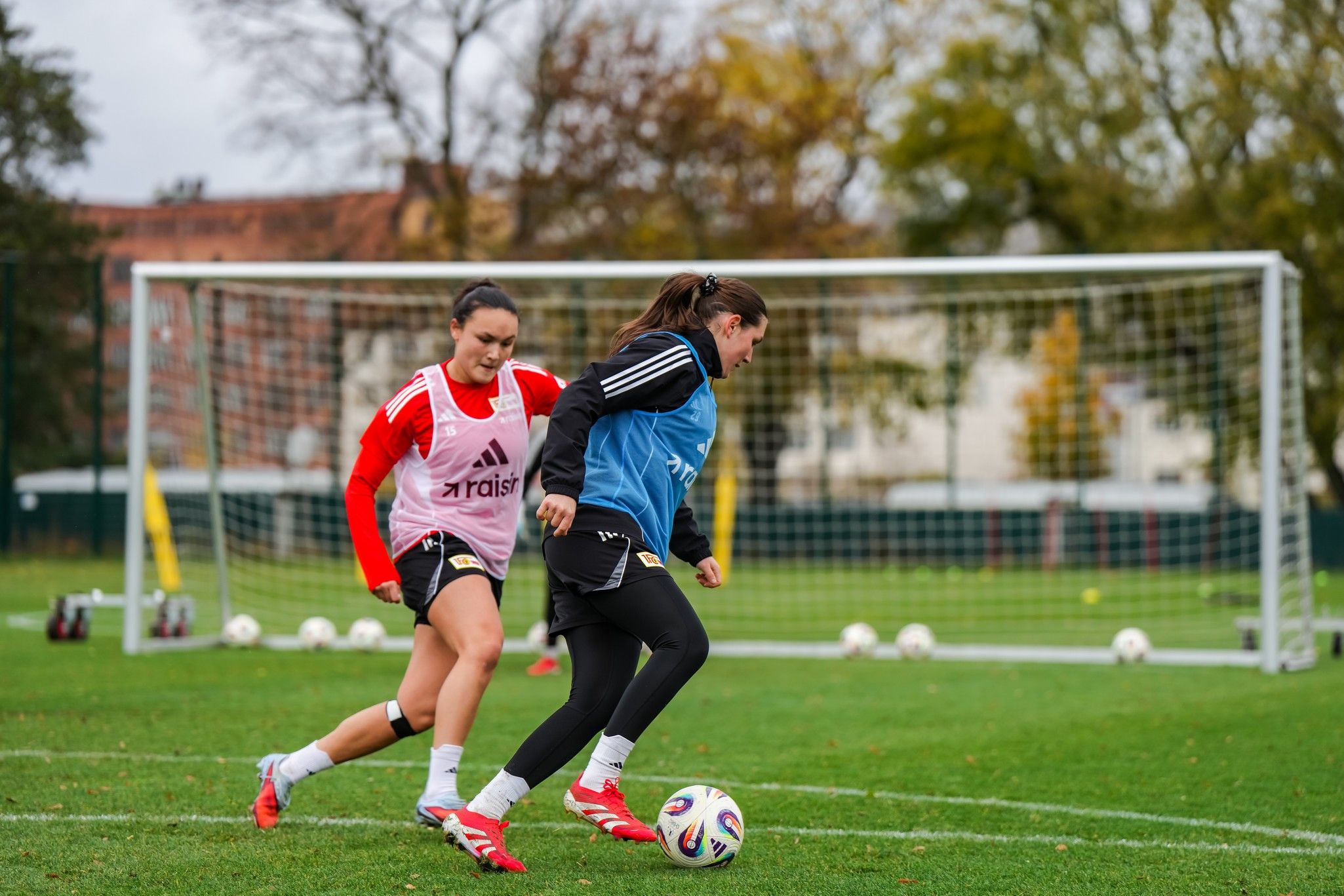 Zwei Fußballspielerinnen in Training, eine dribbelt den Ball, während die andere sie verfolgt. Fußballfeld im Hintergrund.
