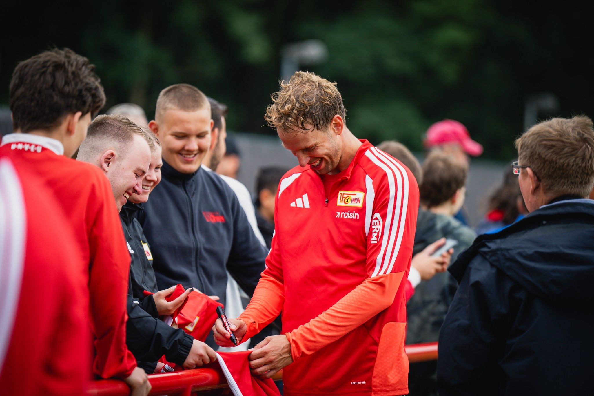 Ein Fußballspieler in einem roten Trikot signiert Autogramme für Fans, die ihn umgeben.