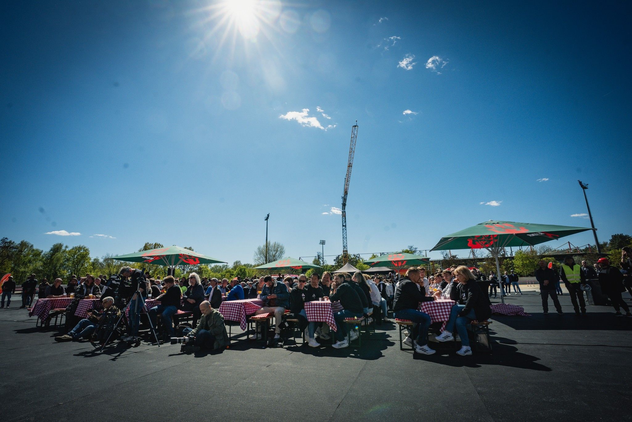 Menschen sitzen an Tischen unter Sonnenschirmen, bei blauem Himmel und Sonnenschein, in einer Außenanlage.