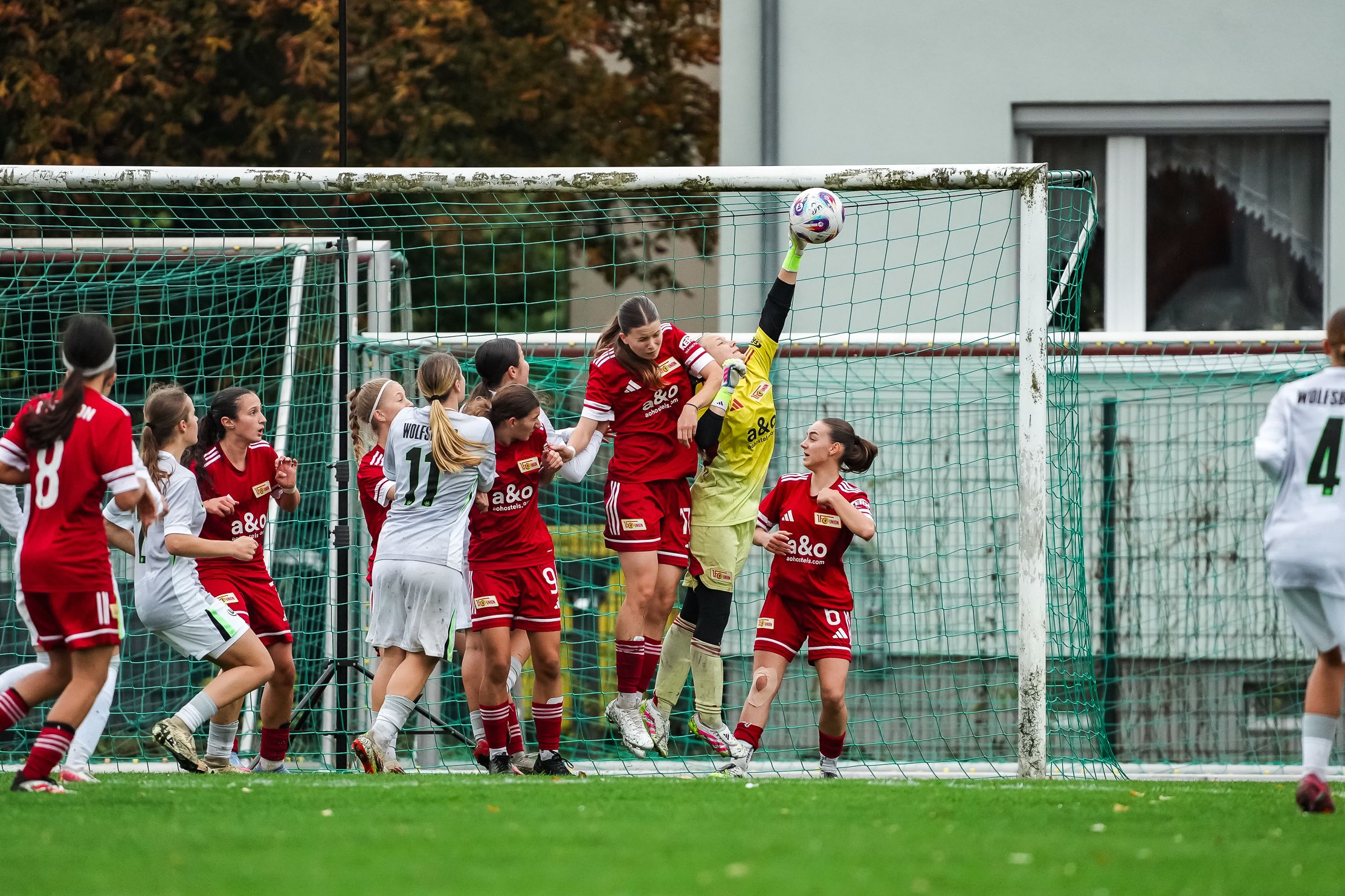 Mädchenfußballspiel: Eine Torwartin springt, um den Ball über das Tor zu halten, während Spielerinnen beider Mannschaften im Abwehrkampf stehen.