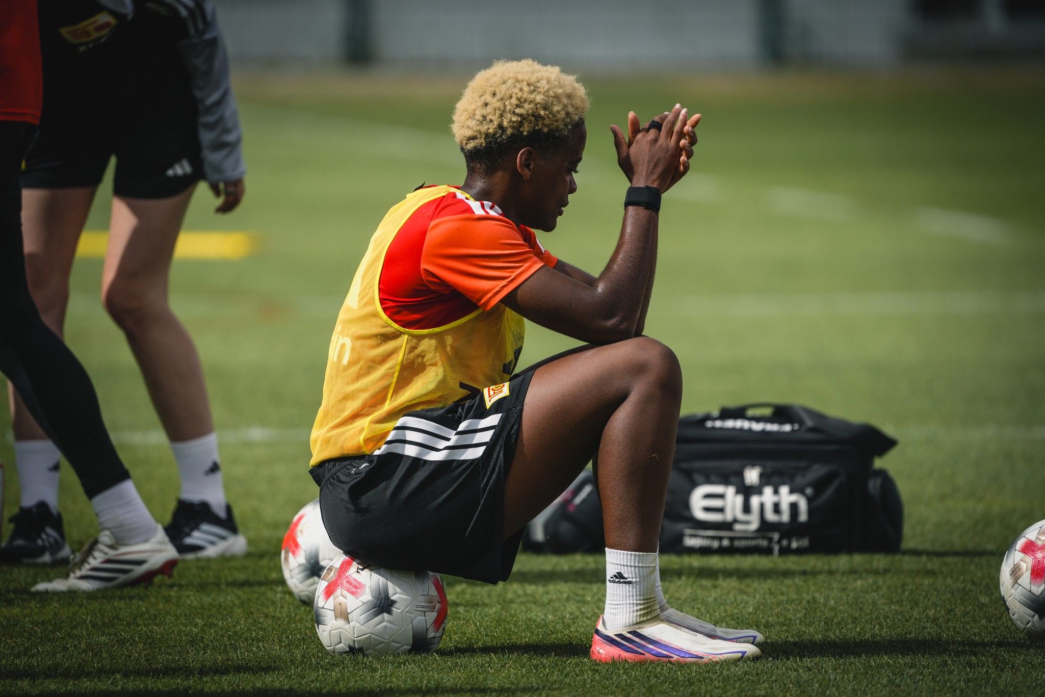 Ein Fußballspieler mit blonden lockigen Haaren sitzt auf dem Rasen, während andere Spieler im Hintergrund trainieren.