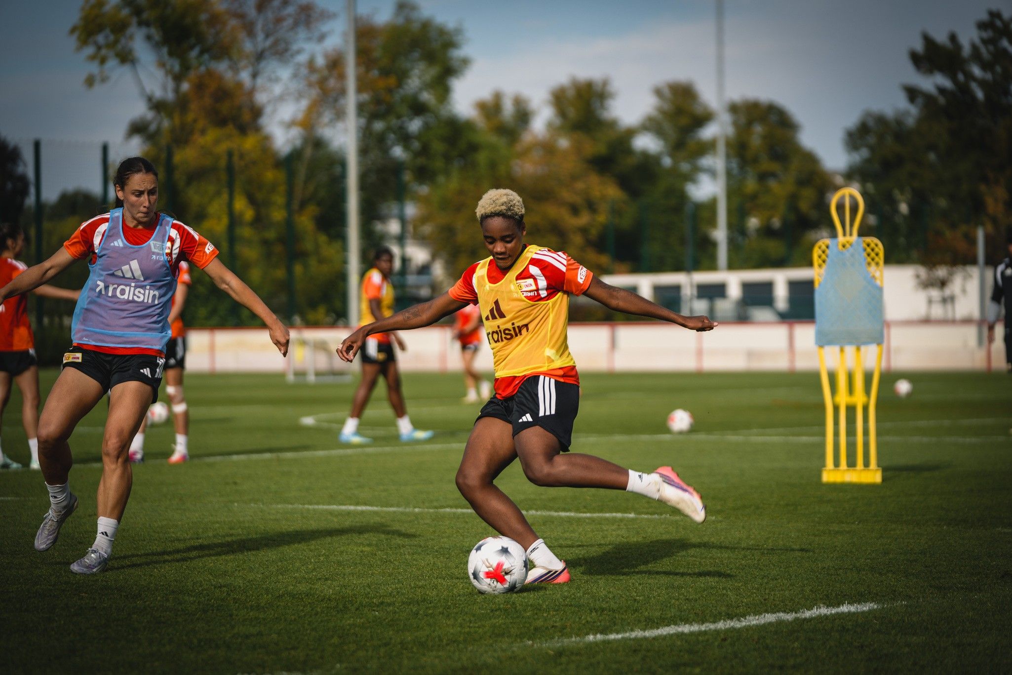 Spielerinnen trainieren auf einem Fußballplatz, während eine Spielerin den Ball mit dem rechten Fuß kickt. Weite Grünflächen und Trainingsausrüstung sind sichtbar.
