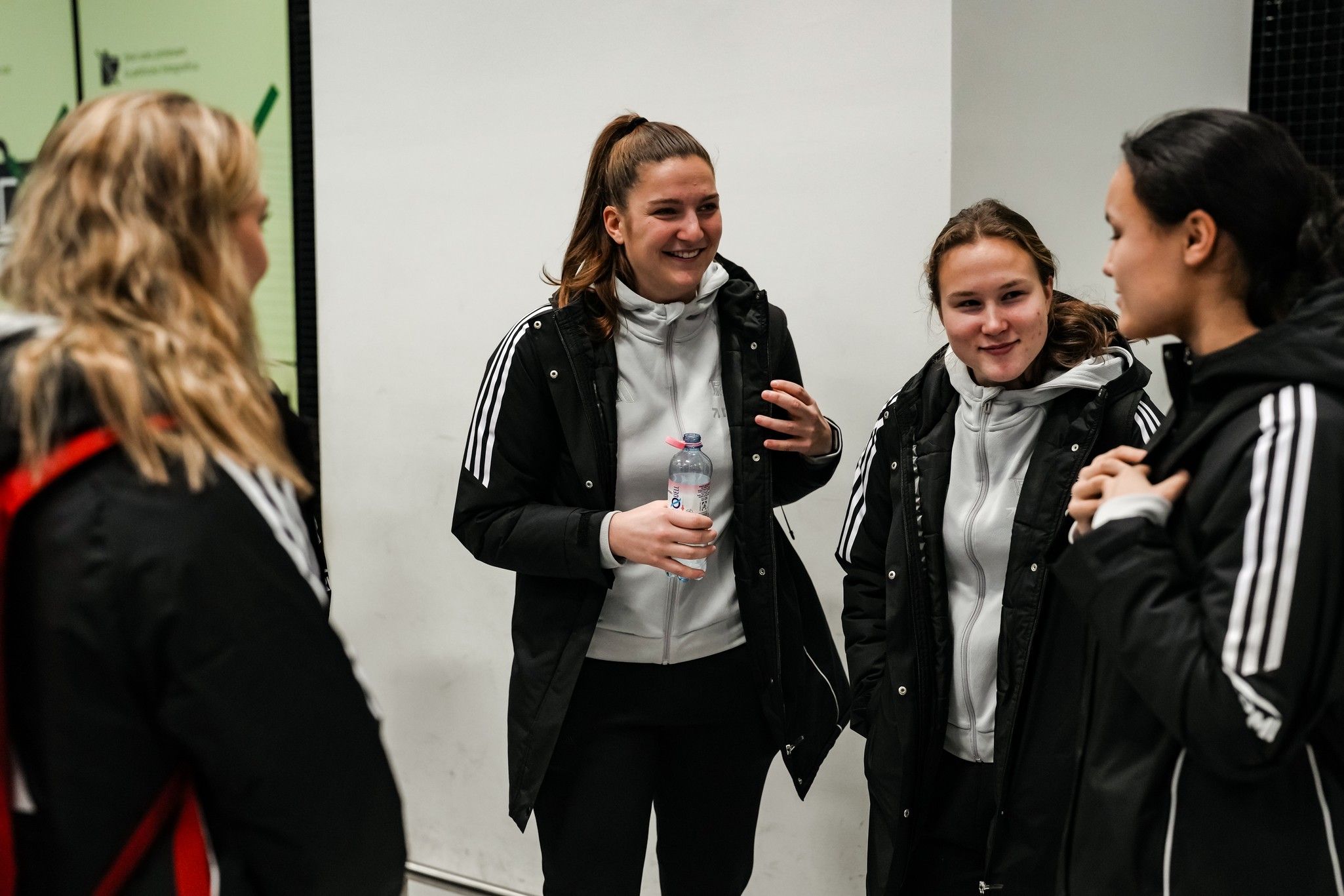 Vier junge Frauen in Sportjacken unterhalten sich lächelnd, eine hält eine Wasserflasche. Hintergrund ist ein heller Raum.