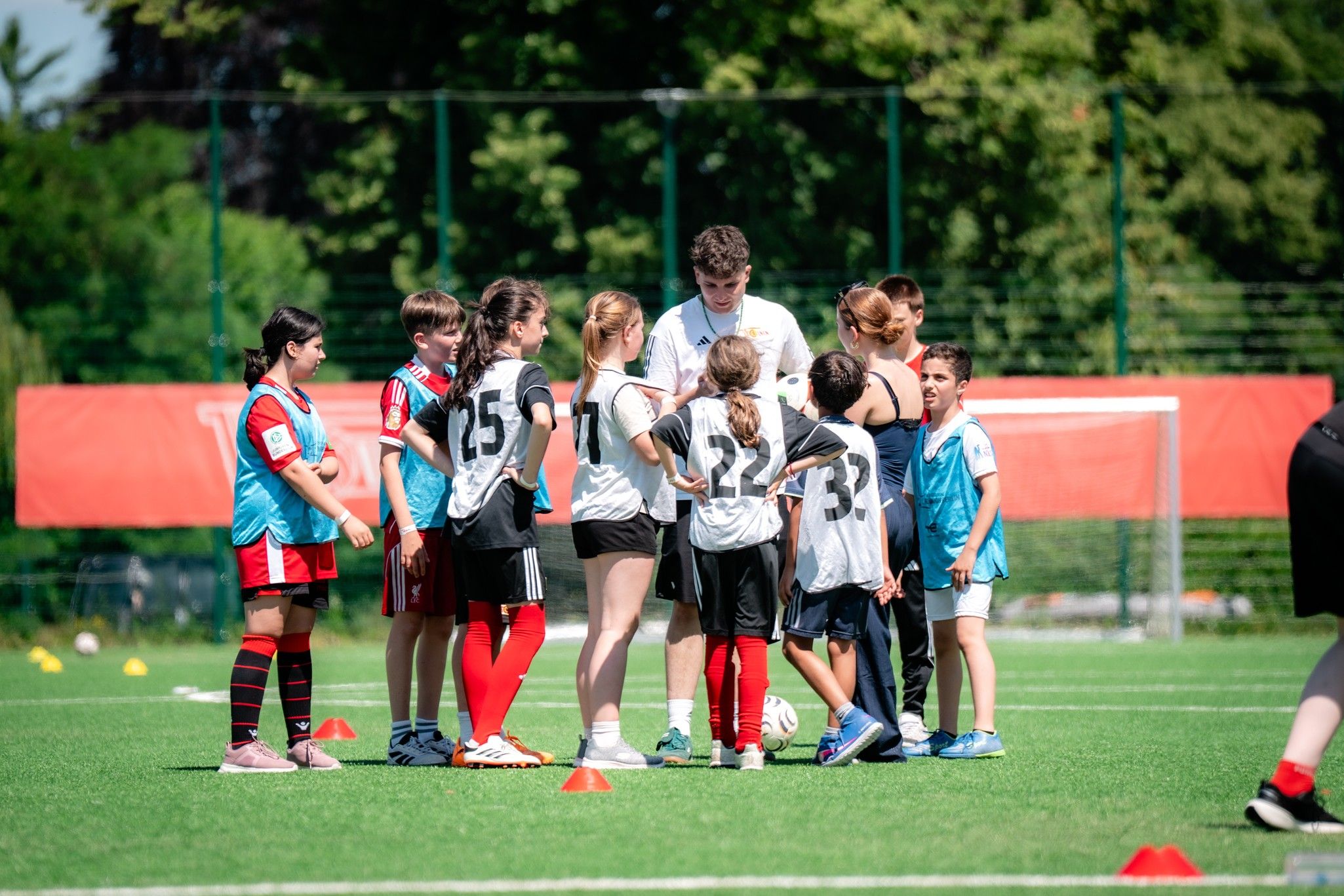 Gruppe von Kindern in Sportkleidung auf einem Fußballplatz, versammelt um einen Trainer in einem weißen Shirt.
