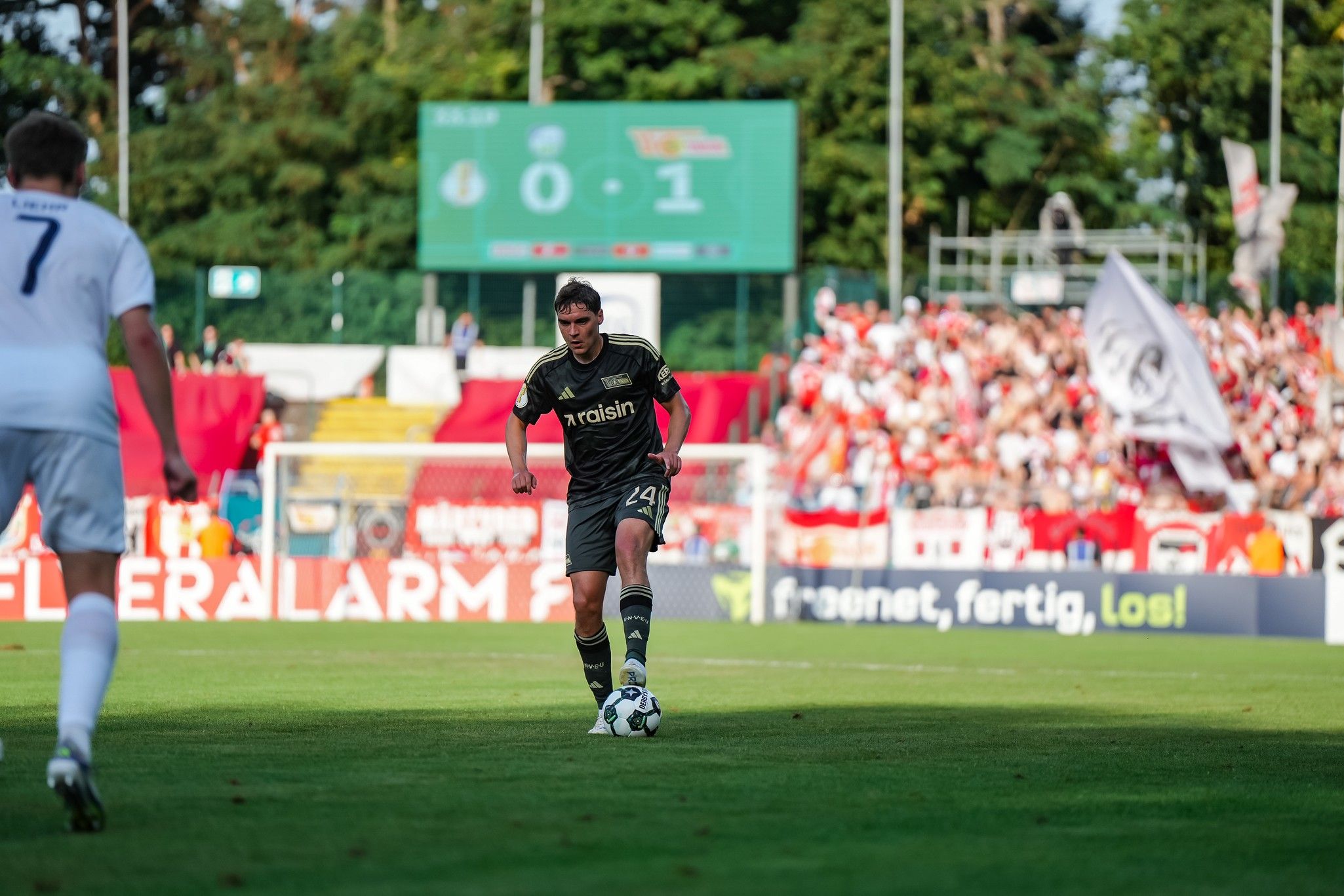 Ein Fußballspieler in dunkler Ausrüstung dribbelt den Ball auf dem Spielfeld, während Zuschauer im Hintergrund jubeln.