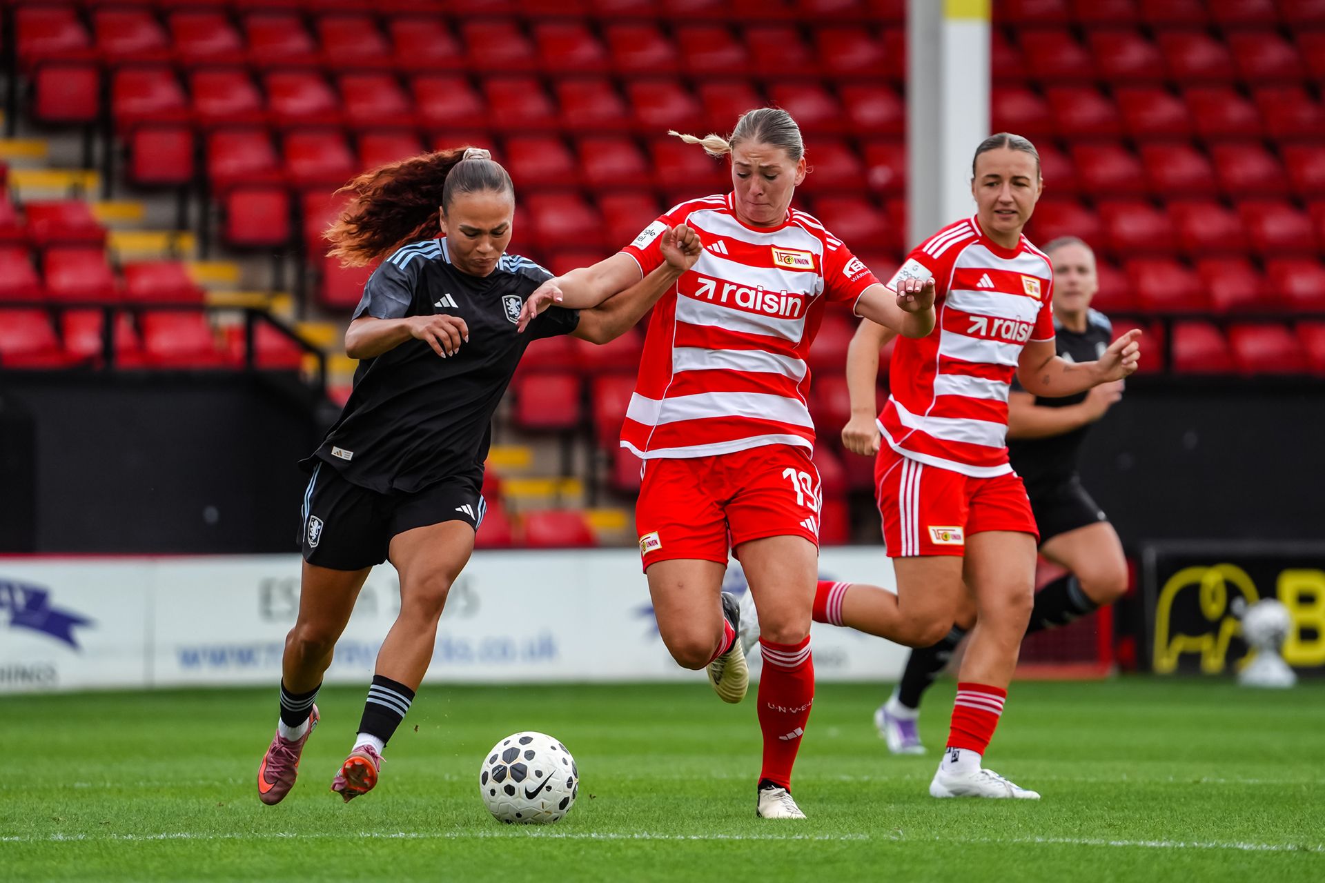 Eine Sportliche Szene im Fußball mit zwei Spielerinnen, die um den Ball kämpfen, während eine dritte Spielerin im Hintergrund läuft.