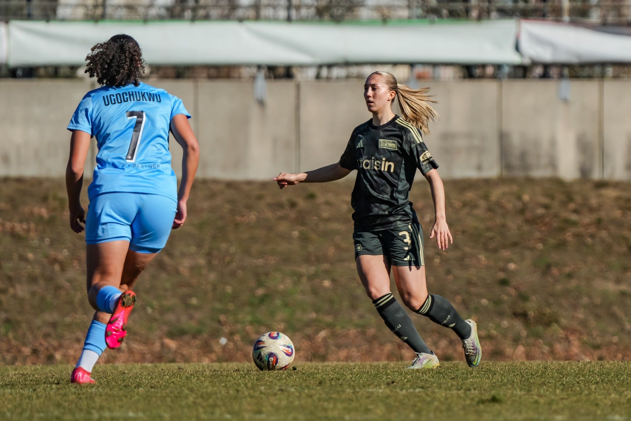 Eine Fußballspielerin in schwarz dribbelt den Ball, während eine Gegnerin in blau sie verfolgt.