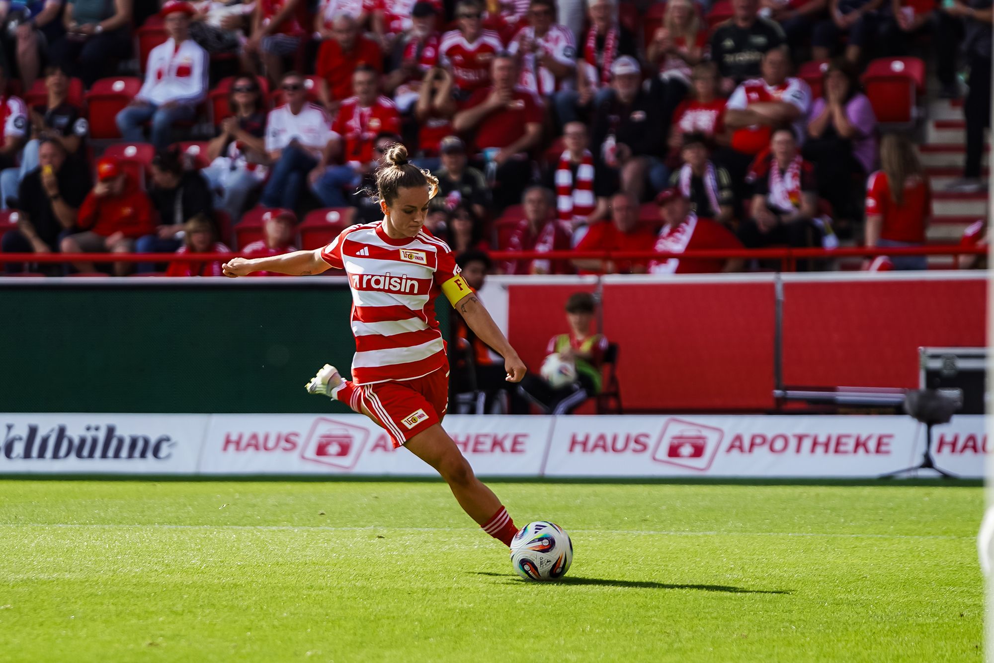 Ein Fußballspieler in rot-weißer Uniform tritt einen Ball auf einem Stadionfeld, umgeben von jubelnden Fans.