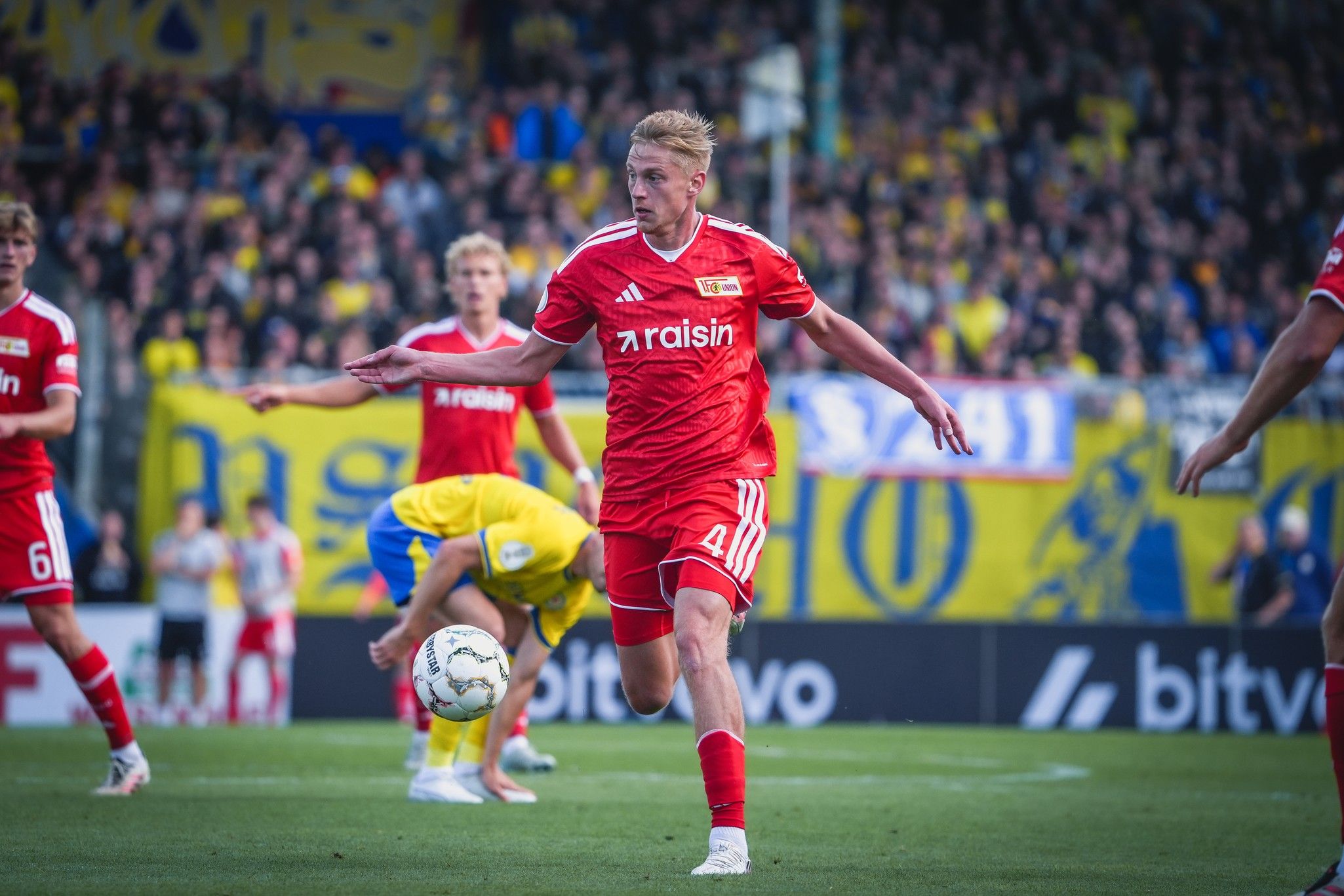 Spieler in roter Fußballuniform läuft mit dem Ball, im Hintergrund jubelnde Fans und eine gelbe Stadionatmosphäre.