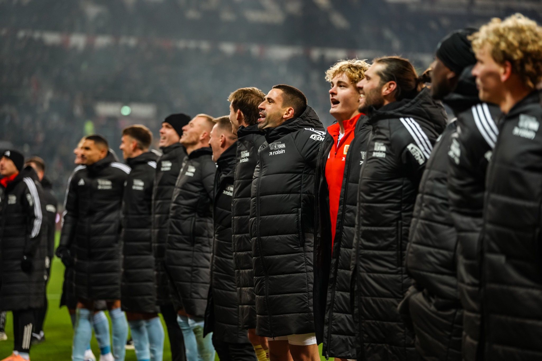 Spieler stehen in schwarzen Winterjacken und singen gemeinsam an der Seitenlinie eines Fußballfeldes.