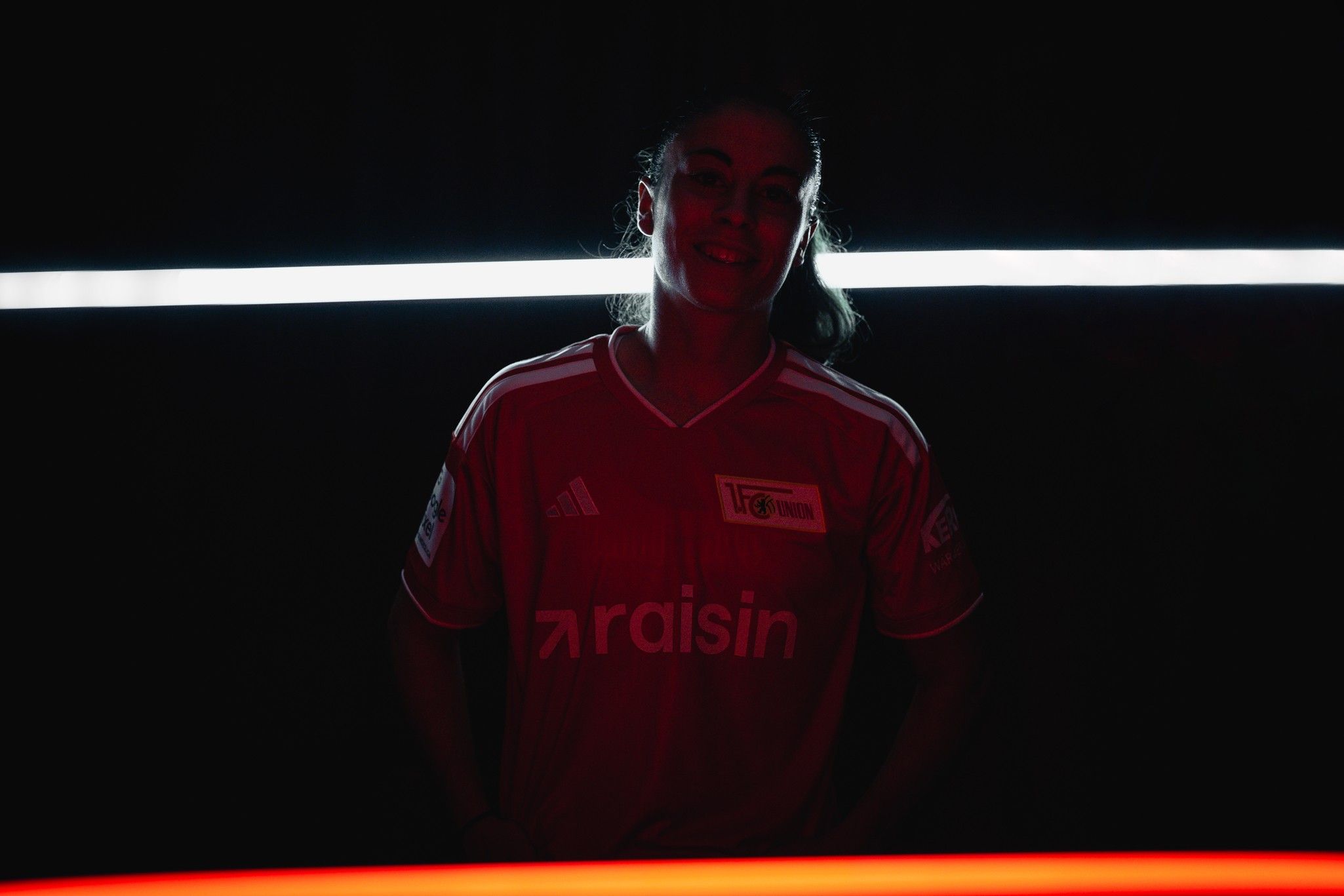 Eine Person in einem rot und schwarz beleuchteten Sporttrikot steht lächelnd vor einem dunklen Hintergrund.