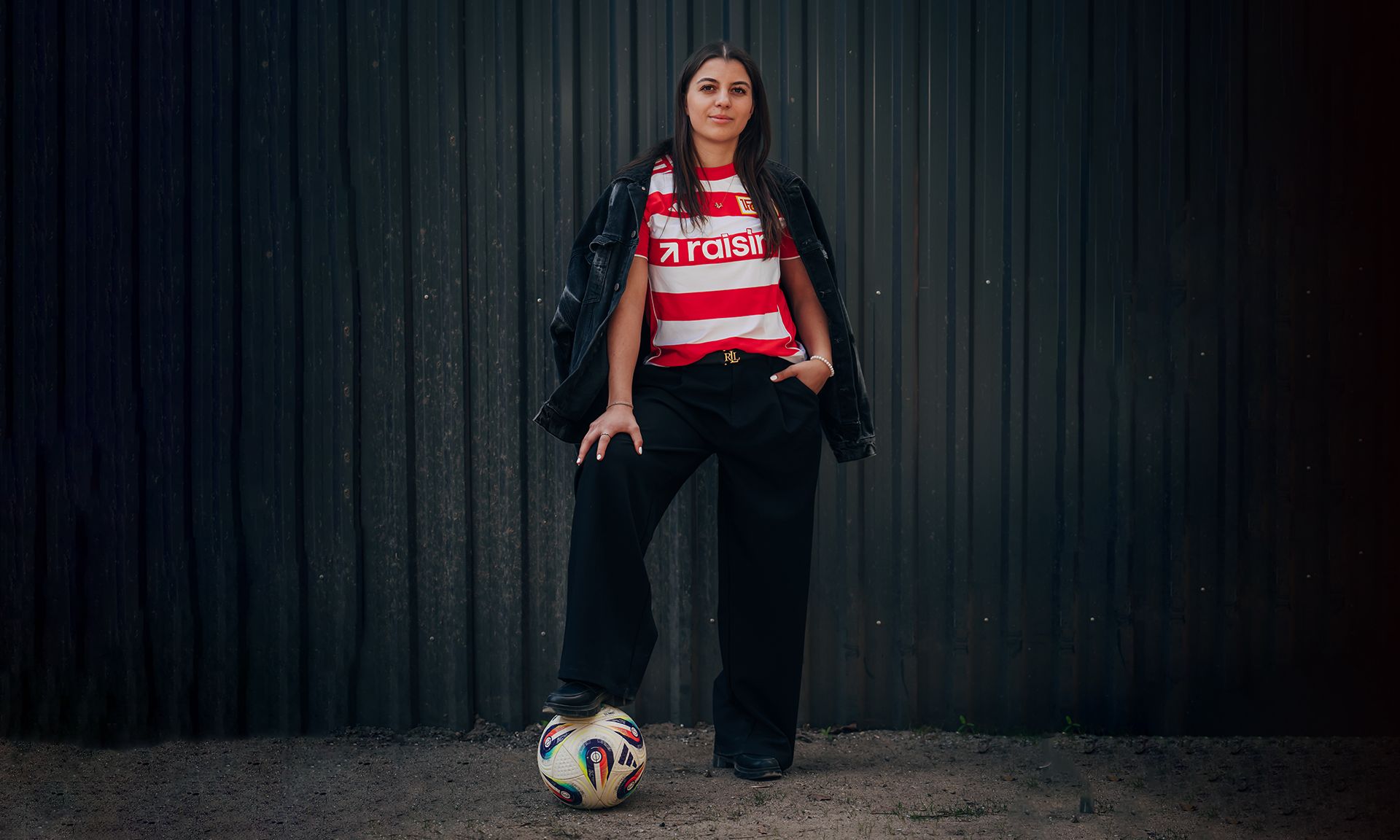 Eine Frau steht mit einem Fuß auf einem Fußball und trägt ein gestreiftes Trikot sowie eine schwarze Jacke vor einem dunklen Hintergrund.