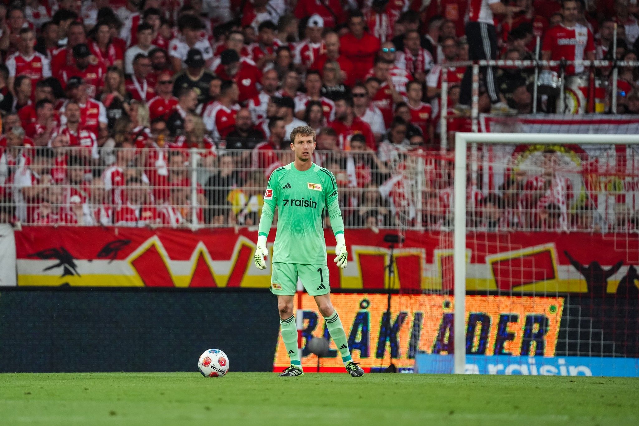 Torwart in grüner Ausrüstung steht im Stadion vor einer jubelnden Menge, mit Torrahmen im Hintergrund.