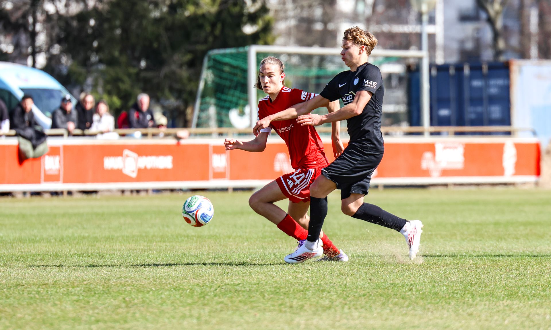 Zwei Fußballspieler in Aktion auf einem grünen Platz, einer im roten, der andere im schwarzen Trikot, während sie um den Ball kämpfen.