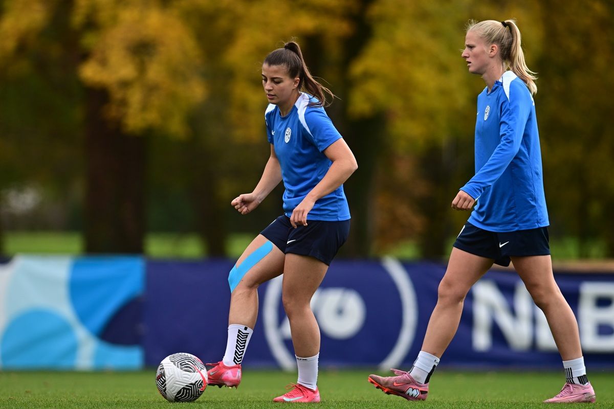 Zwei Fußballspielerinnen in blauen Trikots trainieren auf einem Feld mit einem Ball. Bäume im Hintergrund sind herbstlich gefärbt.
