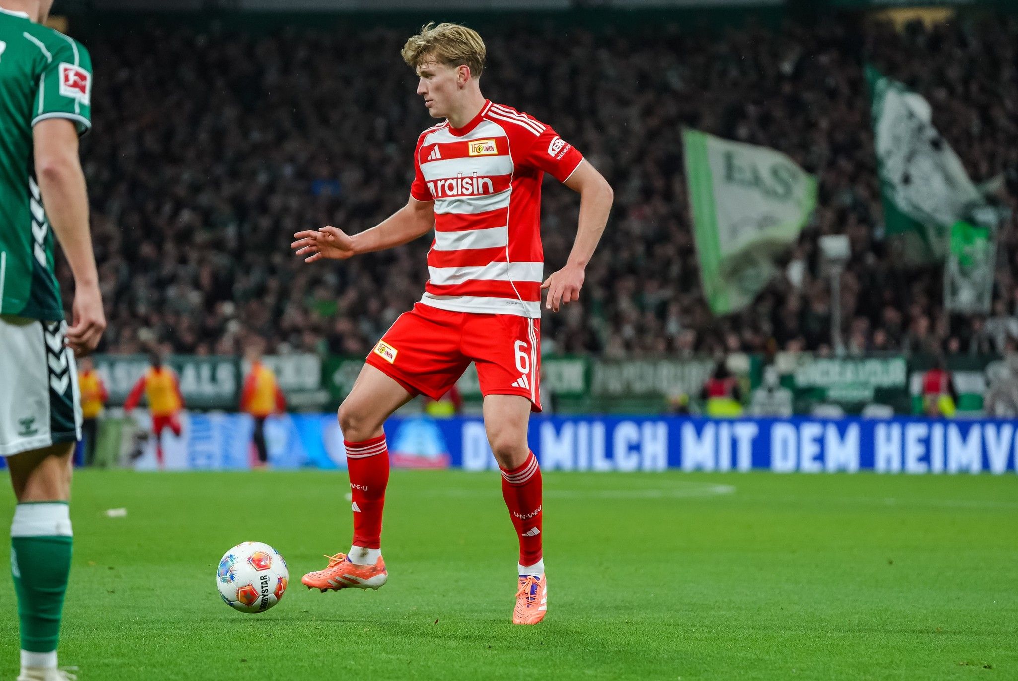 Ein Fußballspieler in gestreiftem Trikot kontrolliert den Ball auf dem Spielfeld, umgeben von jubelnden Fans im Hintergrund.