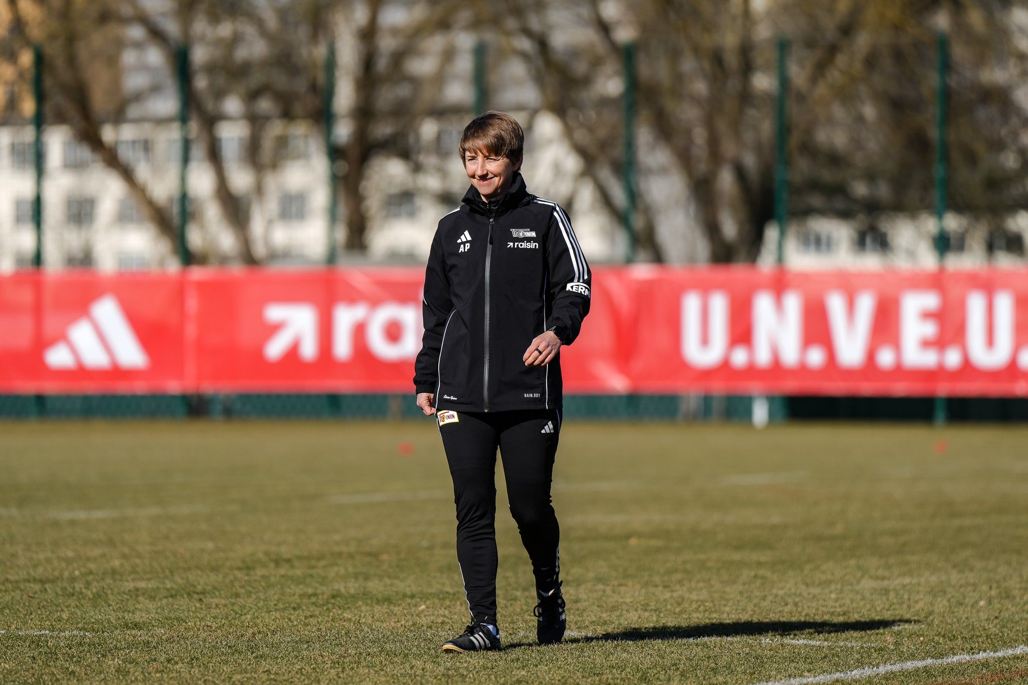 Eine Person in Sportkleidung geht über ein Fußballfeld, im Hintergrund sichtbar ist ein großes Banner.