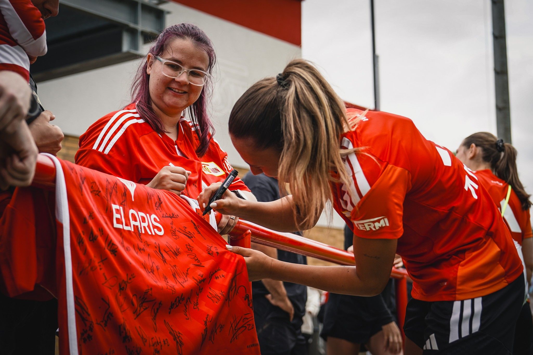 Zwei Frauen in roten Trikots signieren ein Trikot mit vielen Unterschriften im Hintergrund.