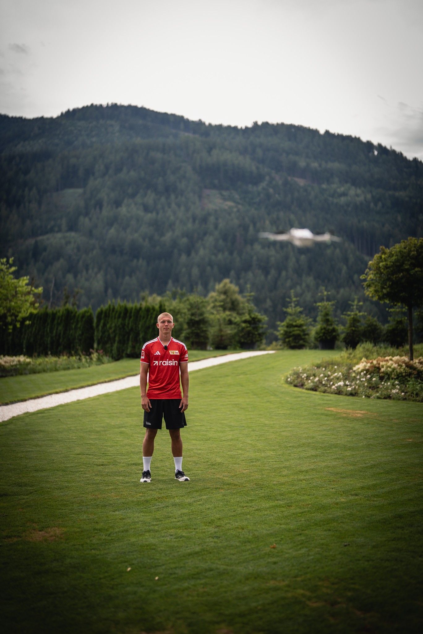 Ein junger Mann in einem roten Sporttrikot steht auf einer grünen Wiese, umgeben von Bäumen und Bergen im Hintergrund.