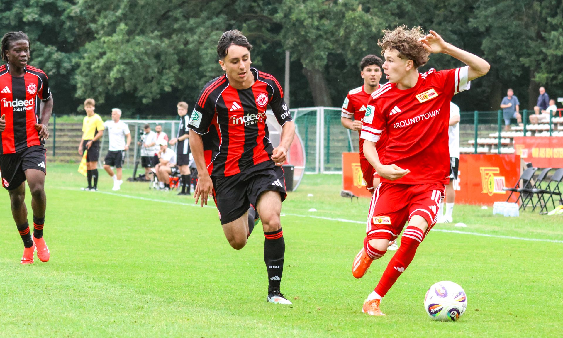 Zwei Fußballspieler auf dem Platz, einer in rotem und der andere in schwarzem Trikot, verfolgen den Ball.