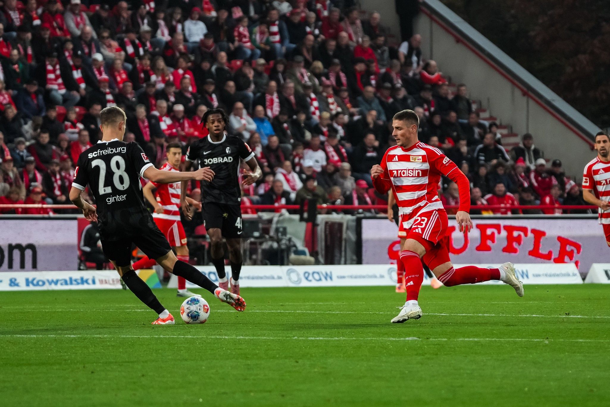 Spielszene im Fußballstadion: Ein Spieler mit der Nummer 28 dribbelt, während ein Gegner mit der Nummer 23 beobachtet. Zuschauer im Hintergrund.