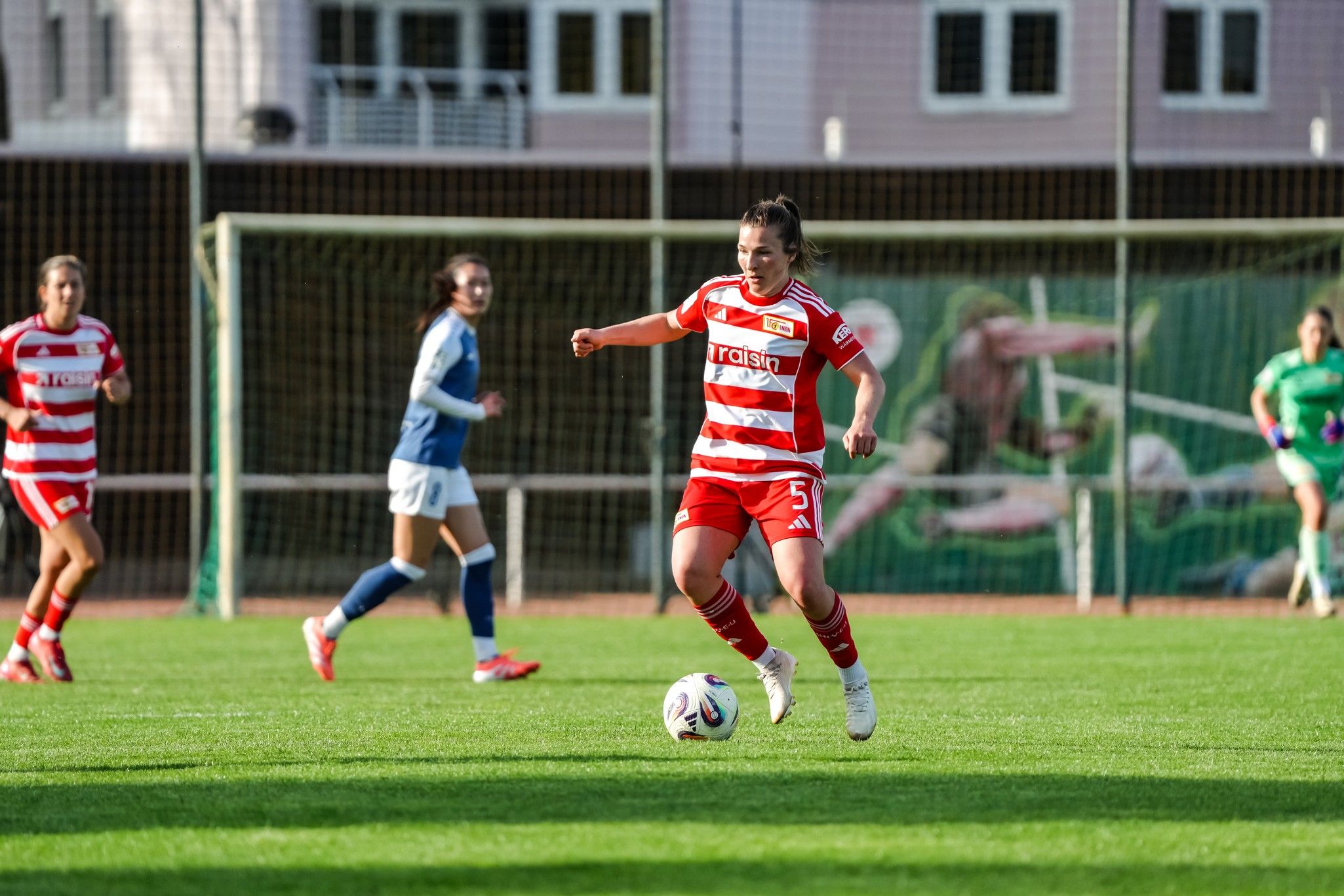 Eine Fußballspielerin in rot-weißem Trikot dribbelt den Ball auf einem Rasenplatz, während Mitspielerinnen im Hintergrund aktiv sind.