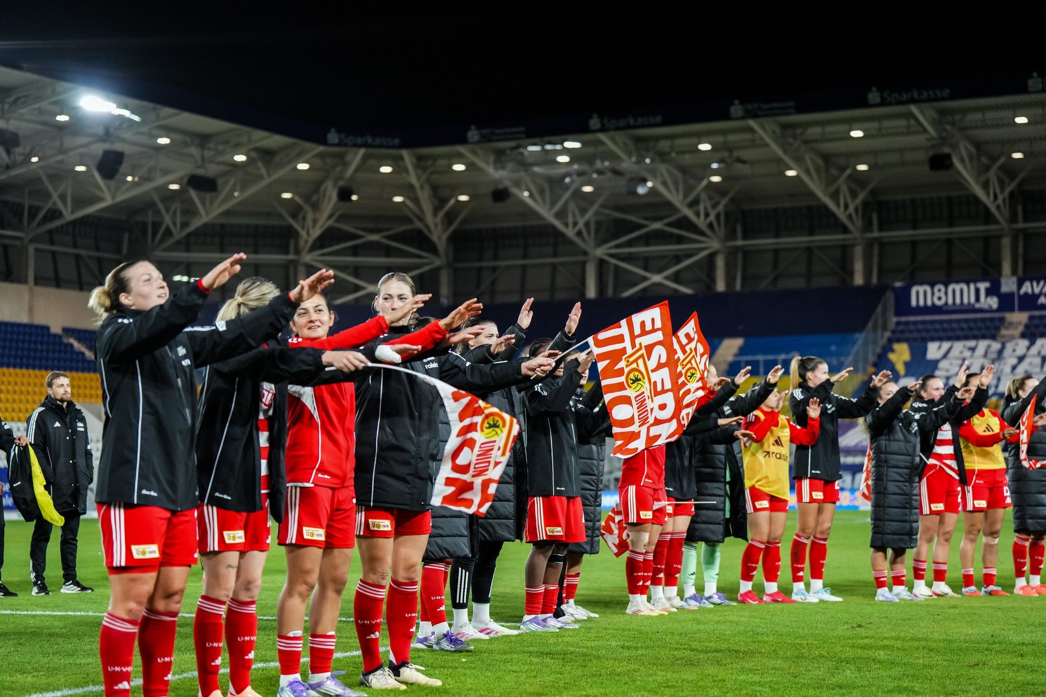 Spielerinnen in roten Trikots stehen auf dem Platz und machen eine Armbewegung, während sie ein Banner halten.