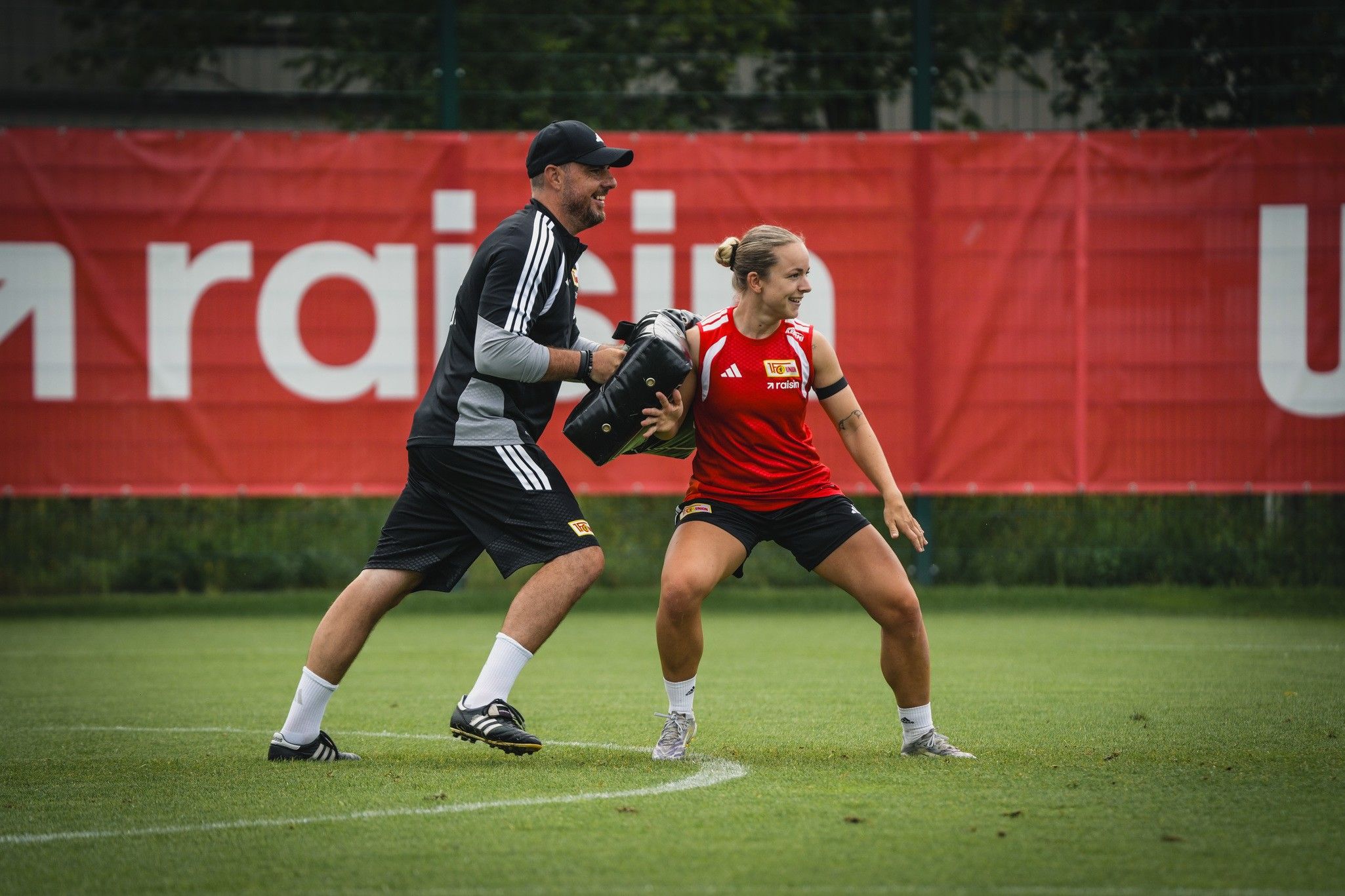Trainingsszene auf einem Fußballplatz: Eine Spielerin übt mit einem Trainer, der ein Polster hält. Rote Wand im Hintergrund.