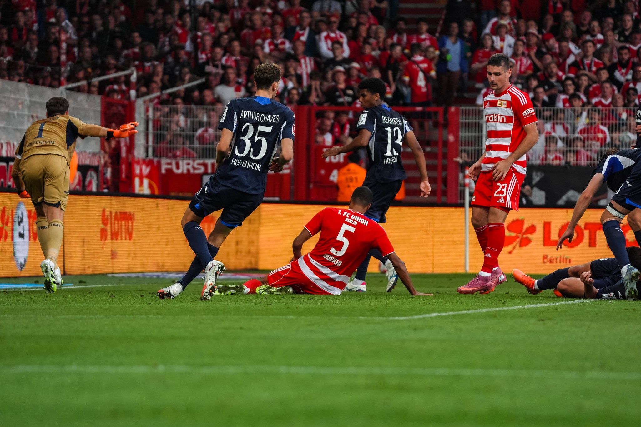 Spielszene im Fußballstadion: Ein Spieler liegt am Boden, während andere Spieler auf dem Spielfeld aktiviert sind. Fans im Hintergrund zu sehen.