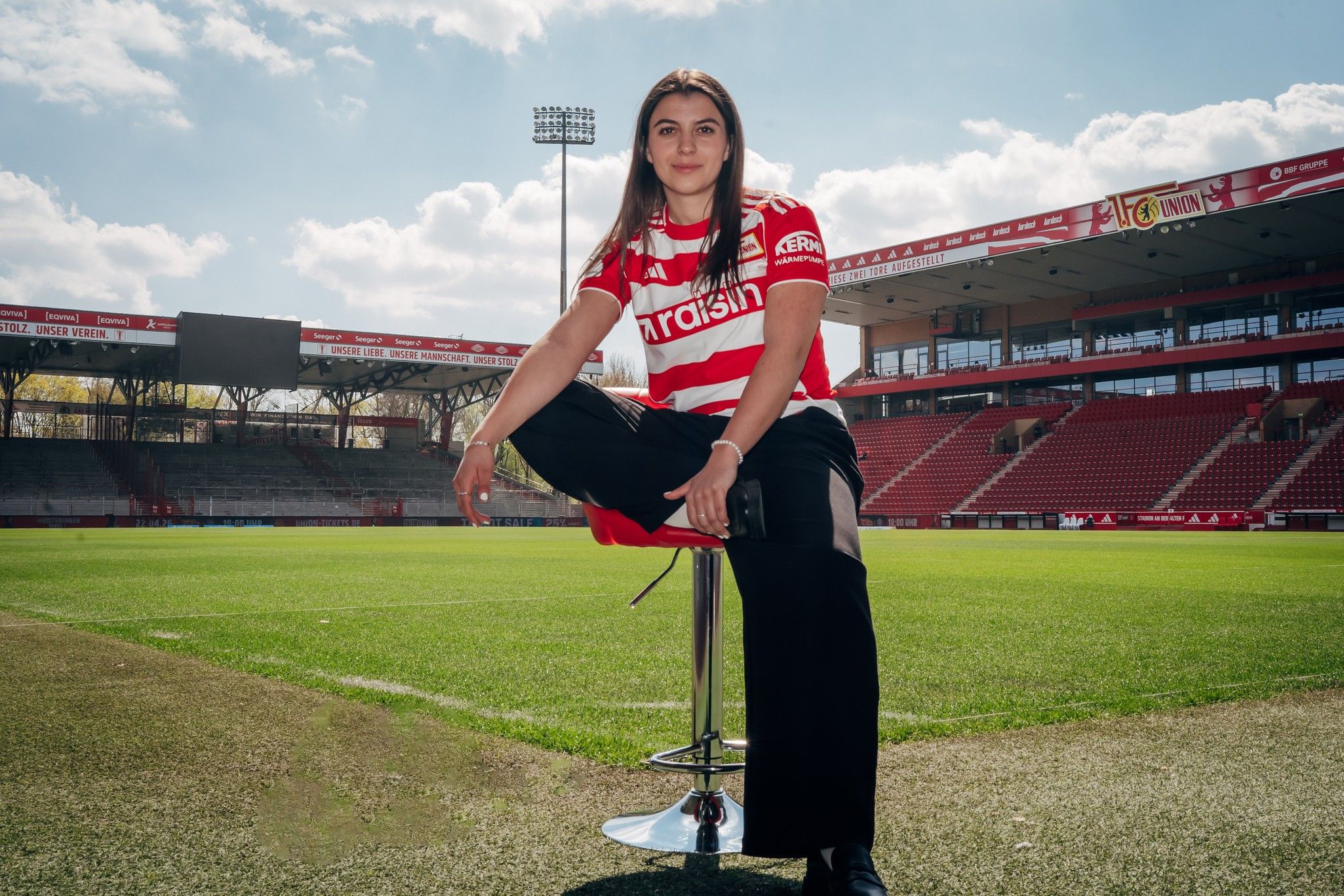 Eine Person sitzt auf einem Hocker, trägt ein rot-weiß gestreiftes Trikot und posiert auf einem Footballfeld mit Stadion im Hintergrund.