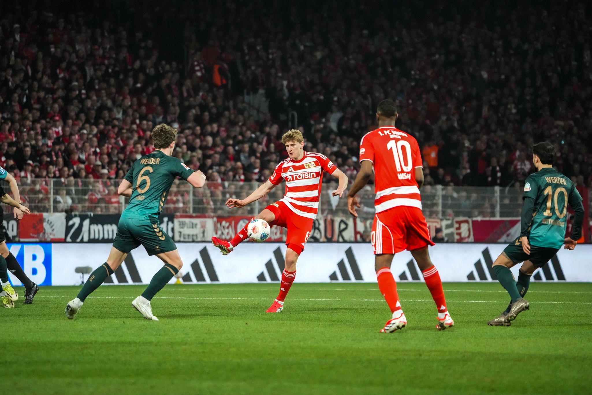 Ein Spieler in rot-weißer Uniform schießt den Ball in einem Fußballspiel, während zwei Gegner in grünem Trikot reagieren.