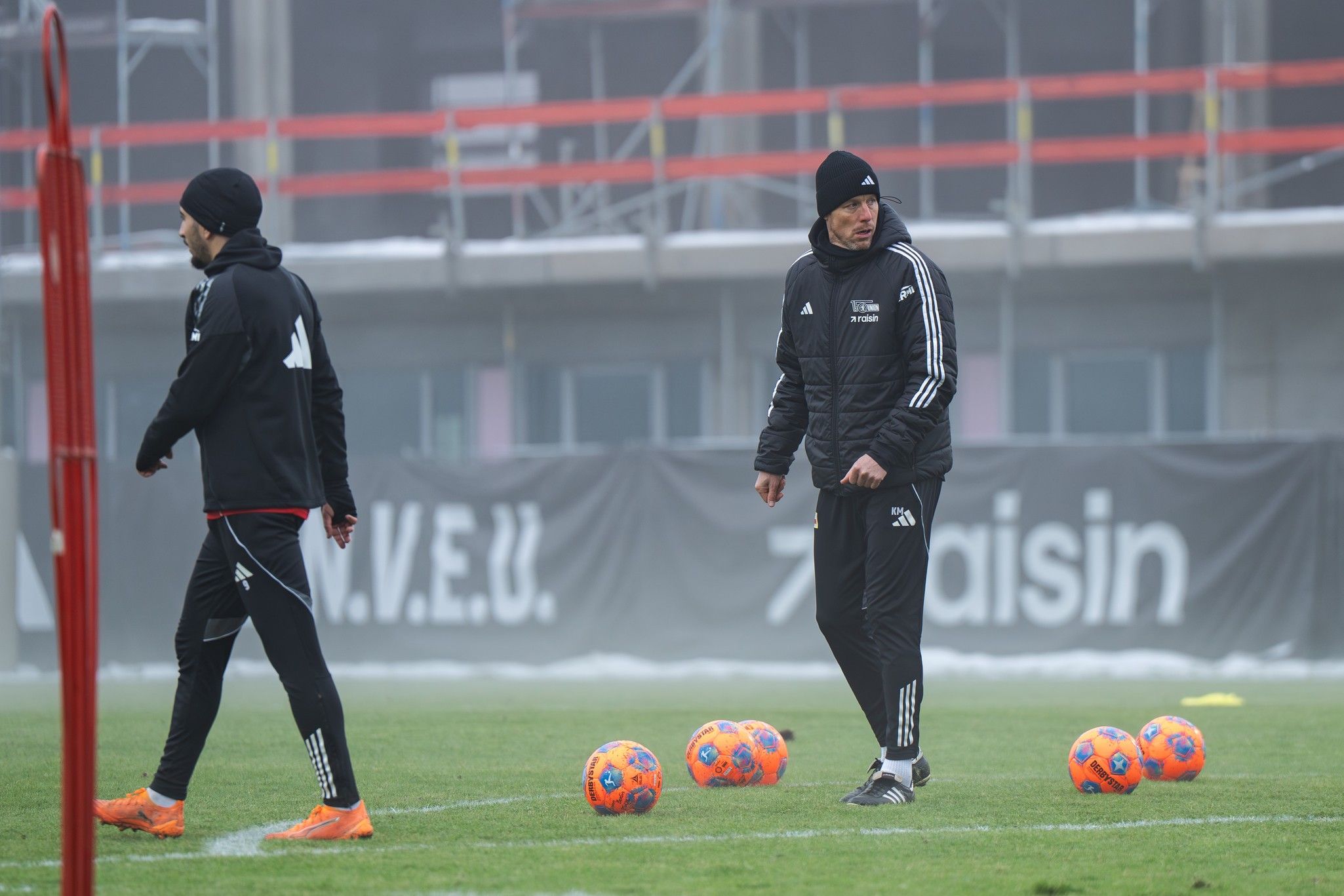 Zwei Personen in Sportkleidung beim Training auf einem Fußballfeld, umgeben von mehreren bunten Fußbällen.