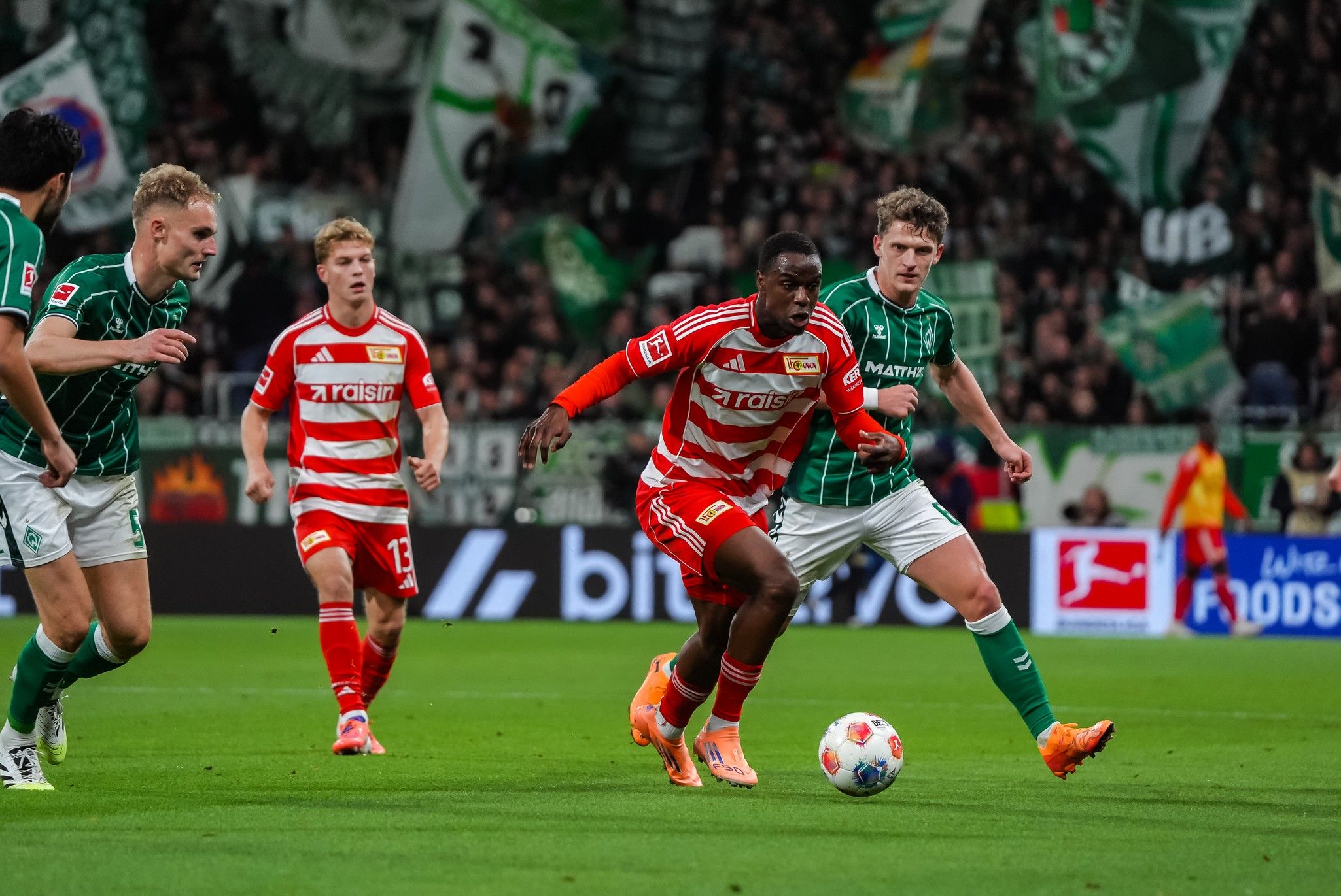 Ein Fußballspieler in rot-gestreiftem Trikot dribbelt den Ball, während zwei Gegner in grünen Trikots versucht, ihn zu stoppen.
