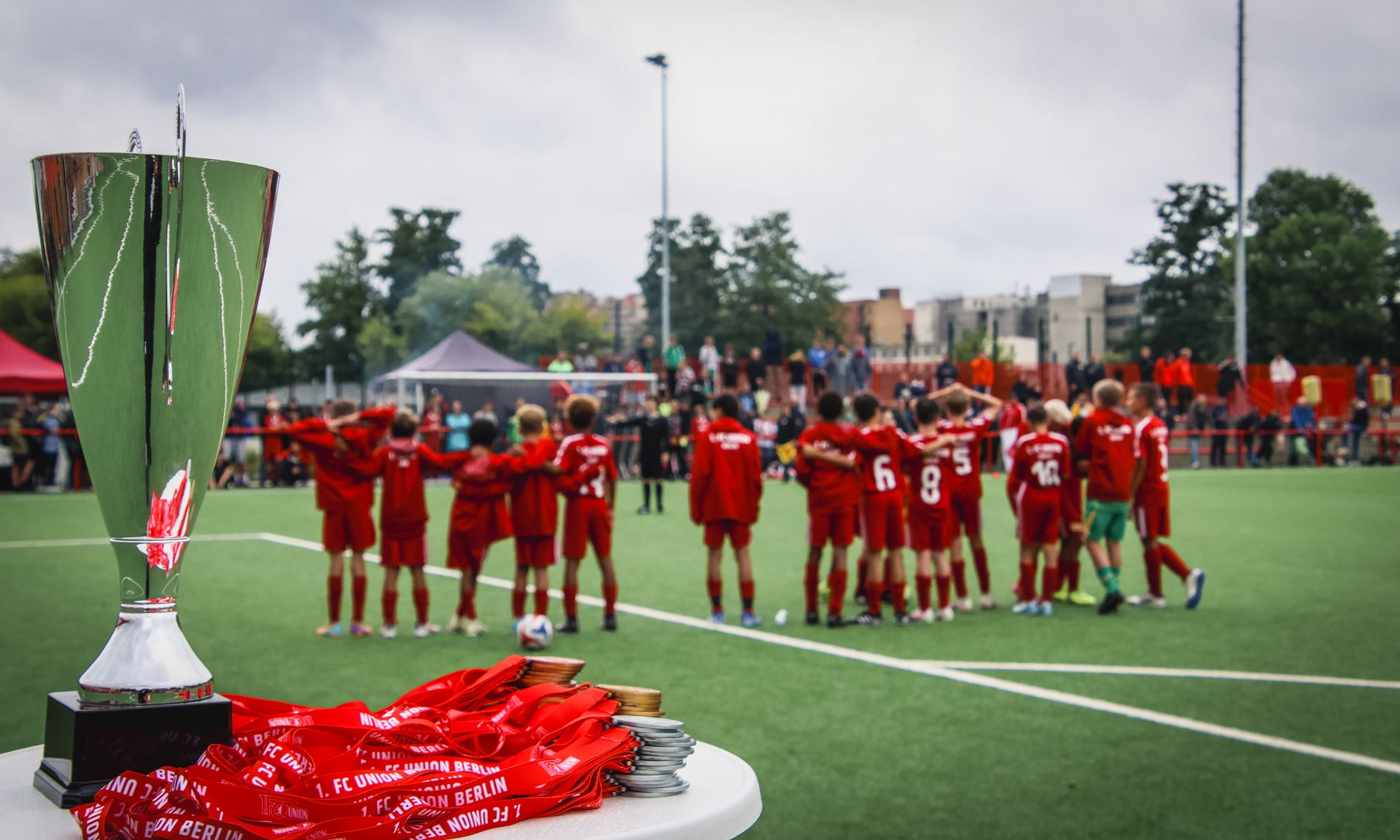 Ein silberner Pokal steht auf einem Tisch, während ein Team in roten Trikots sich zum feiern versammelt.