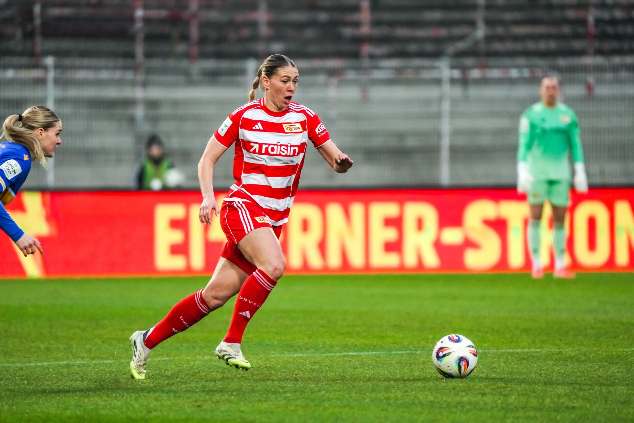 Spielerin in rotem Trikot mit weißem Streifen, läuft dem Ball entgegen. Hintergrund zeigt Stadion und Zuschauer.