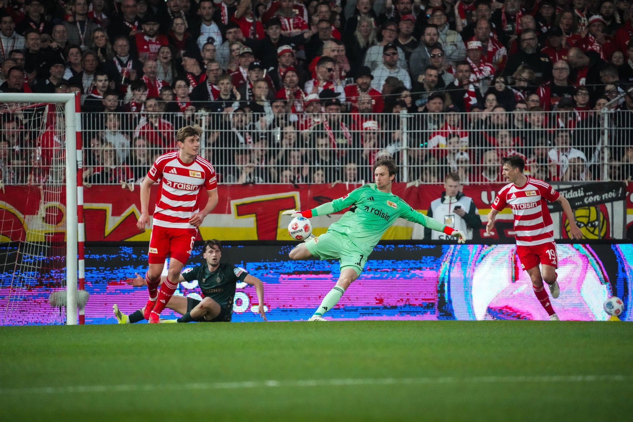 Ein Keeper wehrt einen Schuss ab, während zwei Spieler auf dem Platz reagieren. Viele Zuschauer im Hintergrund.