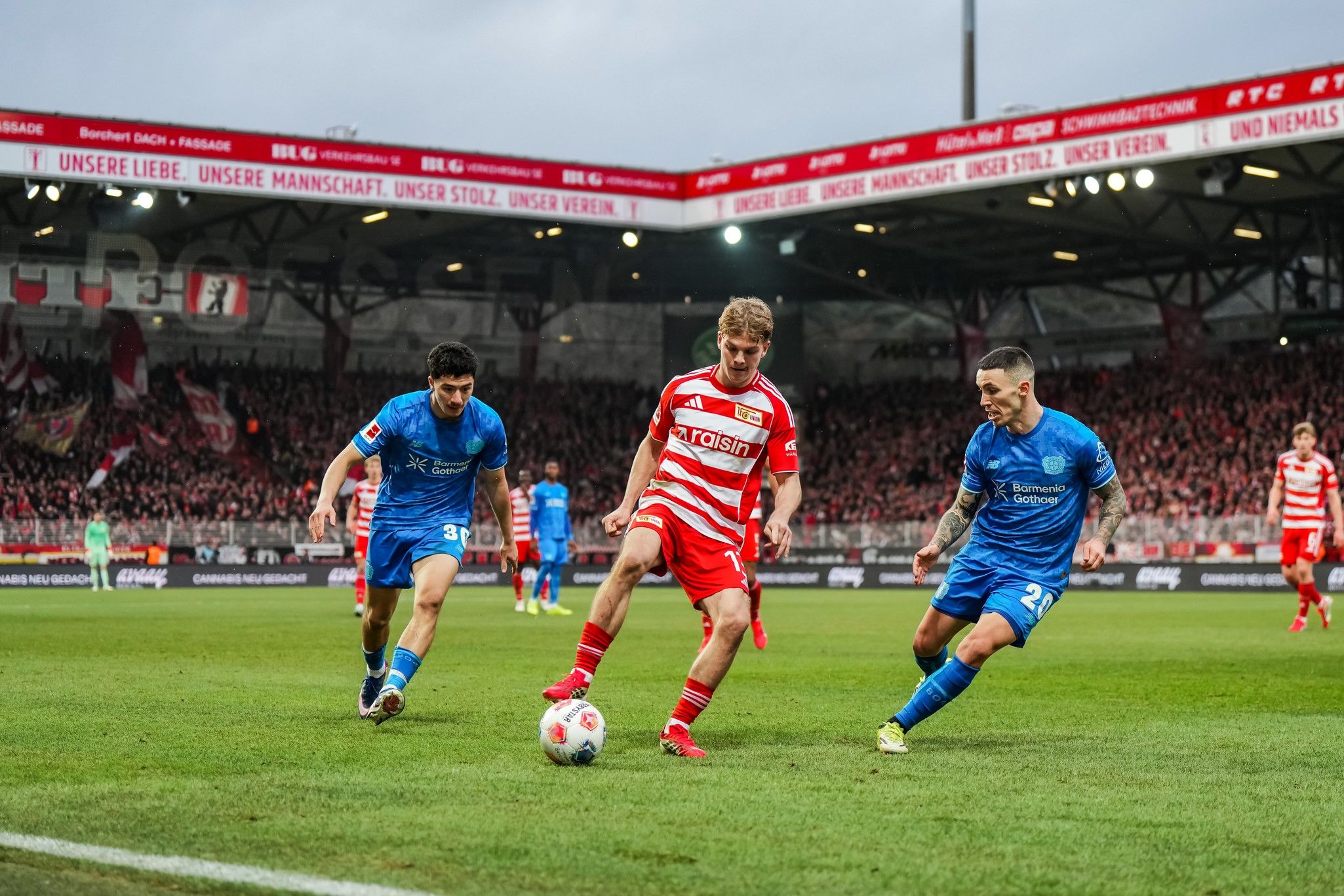 Zwei Spieler in blauen Trikots stehen einem Fußballspieler in rot-weißem Trikot gegenüber, während sie um den Ball kämpfen. Stadion im Hintergrund.