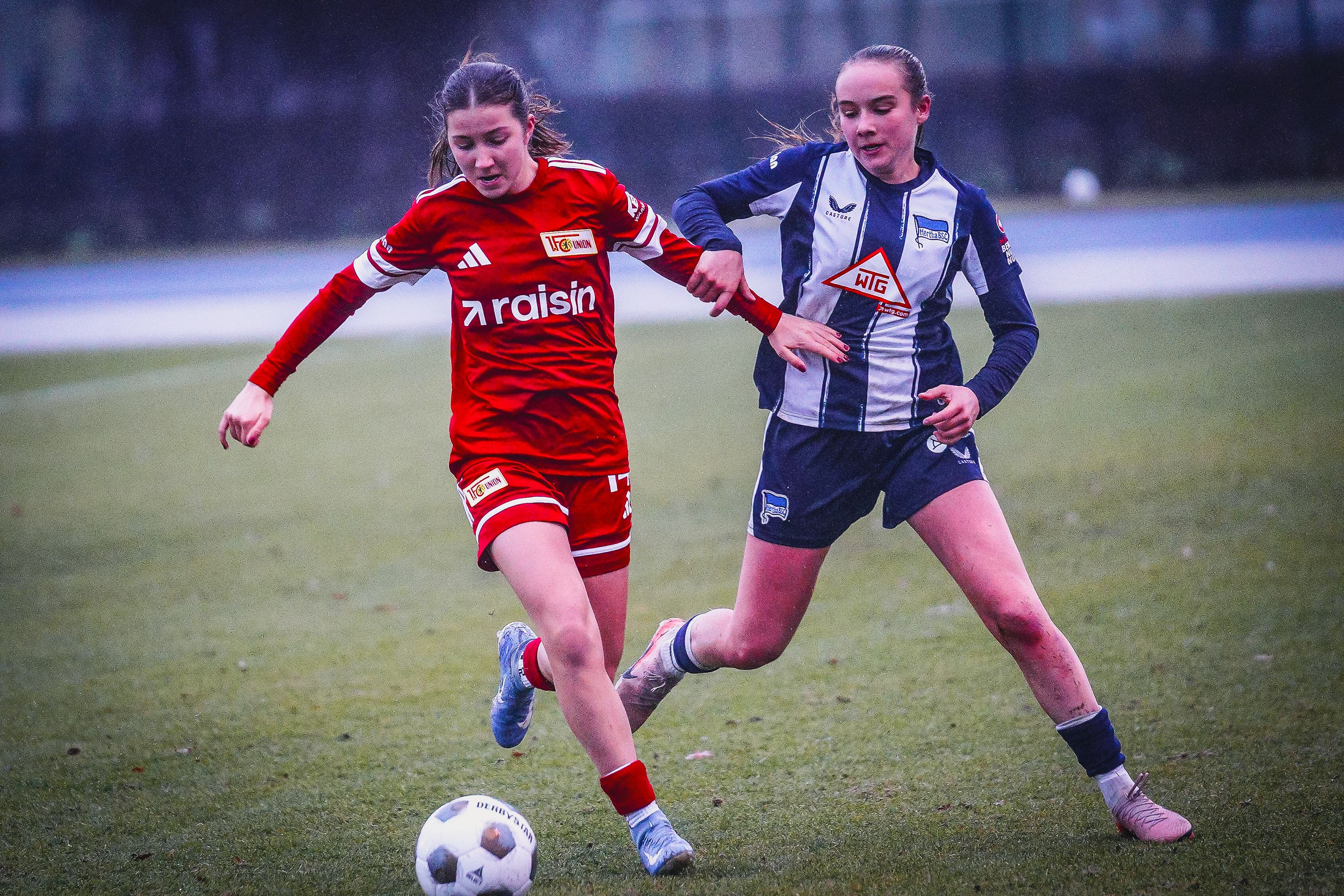 Zwei junge Fußballspielerinnen in Aktion auf einem Fußballfeld, eine in rotem, die andere in dunkelblau-weißem Trikot, beide verfolgen den Ball.