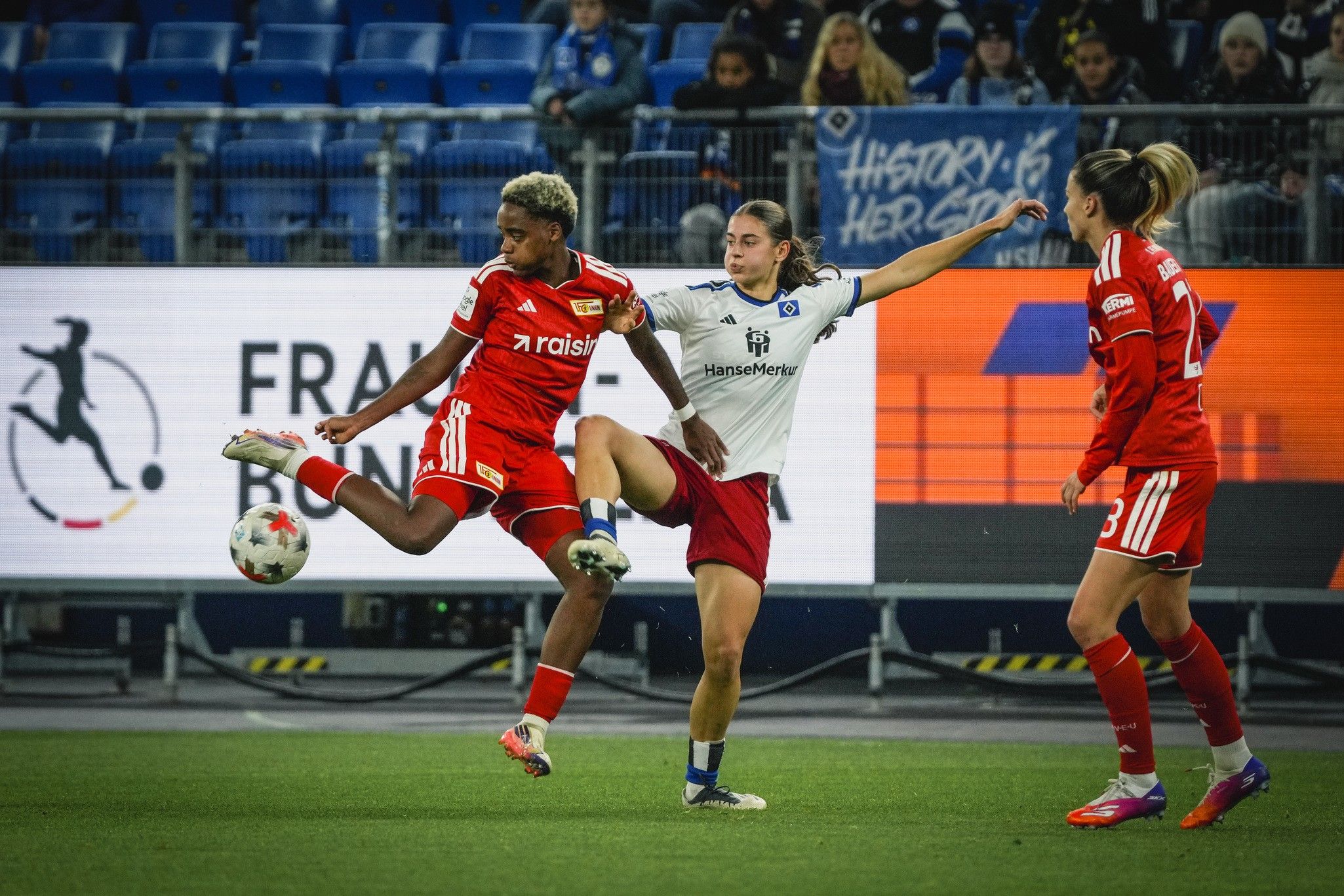 Eine Fußballspielszene mit zwei Spielerinnen im Zweikampf, während eine dritte Spielerin im Hintergrund schaut.
