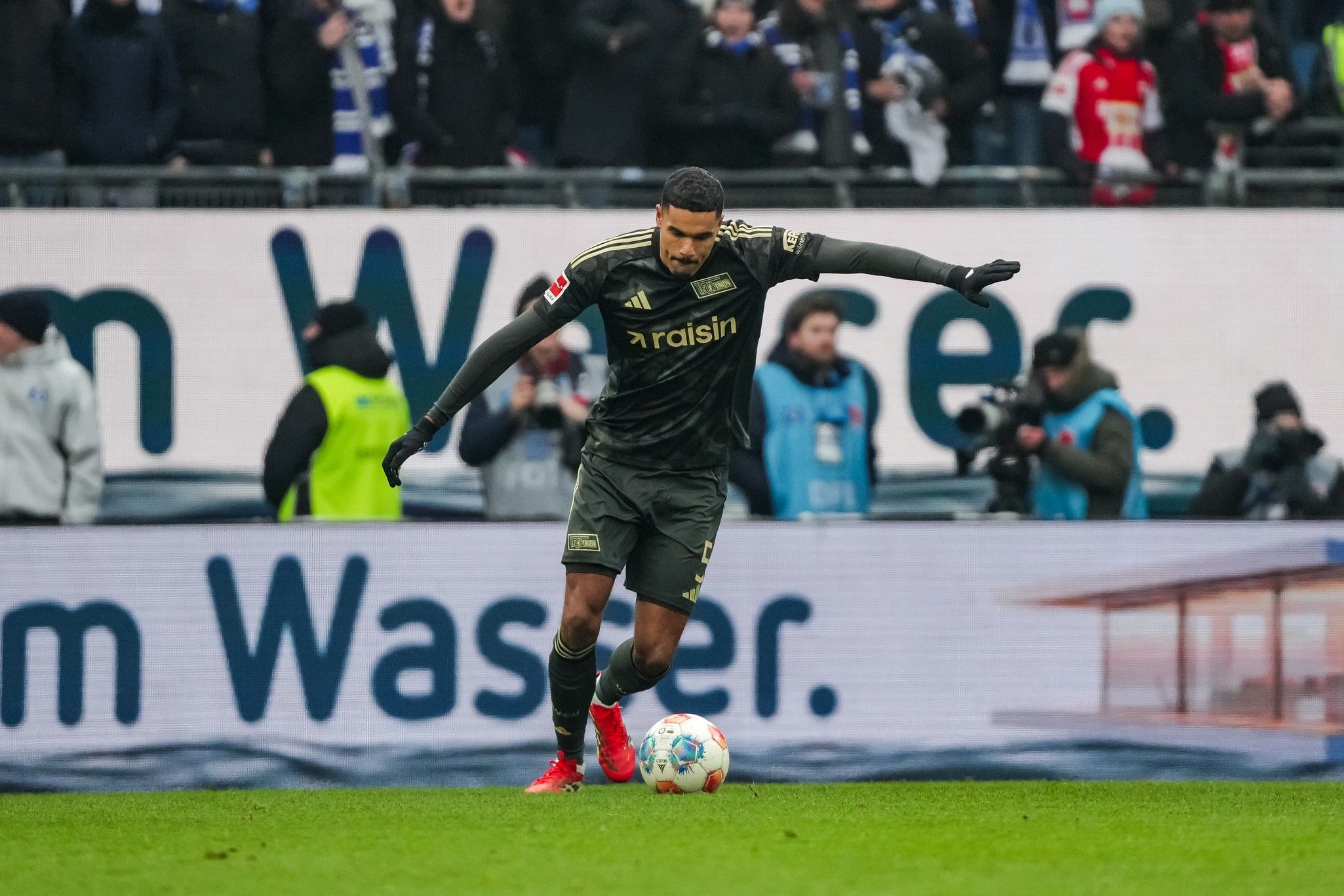Ein Fußballspieler in schwarzem Trikot führt einen Freistoß aus, während Zuschauer im Hintergrund stehen.
