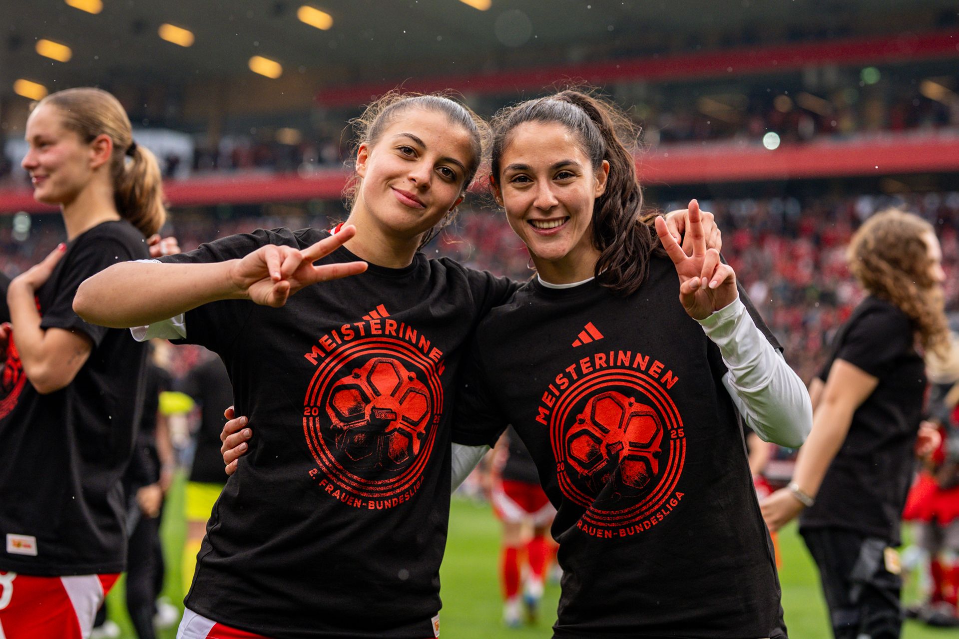 Zwei Frauen in schwarz-gelben T-Shirts posieren lächelnd mit Peace-Zeichen nach einem Fußballspiel.