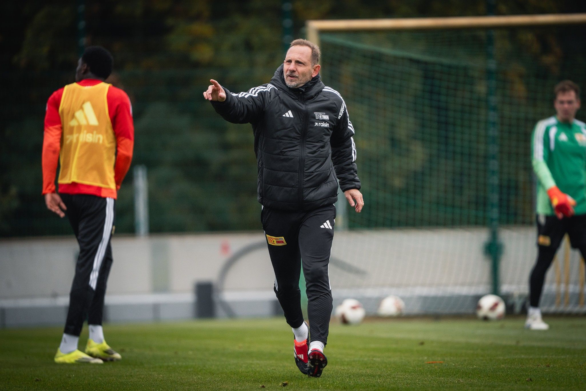 Trainer gibt Anweisungen während eines Fußballtrainings, Spieler im Hintergrund hören aufmerksam zu.